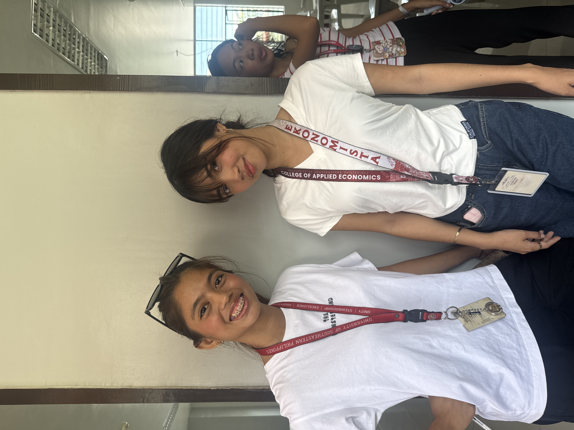Two young women stand in front of a mirror. The woman in the lower portion of the image is smiling, wearing a white t-shirt, with sunglasses propped on her head. She has a lanyard around her neck that reads "UNIVERSITY OF SOUTHEASTERN PHILIPPINES". A second woman, also smiling, is positioned above her. She is wearing a white t-shirt and a lanyard that reads "UNIVERSITY OF SOUTHEASTERN PHILIPPINES COLLEGE OF APPLIED ECONOMICS". The top portion of the image shows the reflection of a third person, also wearing a white shirt, lying down. A window with bars is visible in the reflection. The lanyards suggest the location is in Davao City, Philippines.