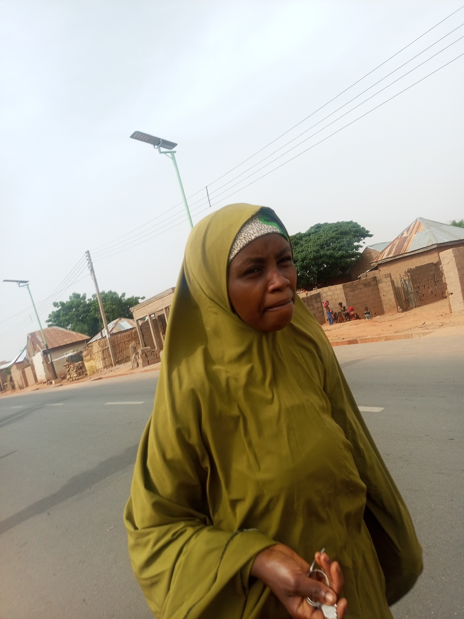 A medium close-up shot captures a woman in a green hijab/khimar standing on the edge of an asphalt road, looking slightly to her right. Her dark skin is visible on her face and hands. She holds a set of keys in her right hand, which is lightly moisturized. Her expression appears neutral or slightly pensive.
The setting is a street in Tempure, Nigeria, characterized by an overcast or hazy sky. The road features a white dividing line. In the background, several single-story structures constructed from brick and mud are visible, many topped with corrugated metal roofs. A prominent green tree stands in the mid-ground, providing a focal point amidst the buildings. On the right side of the background, a small group of figures, likely children, are gathered near one of the structures.
Two light-green poles with solar-powered streetlights are seen against the light sky. Overhead, multiple thin utility lines stretch across the frame. The overall atmosphere suggests a typical daytime scene in a semi-rural or developing urban environment, with the soft, diffused light indicating either an overcast day or a time of day with indirect sunlight. There is no visible text in the image.
