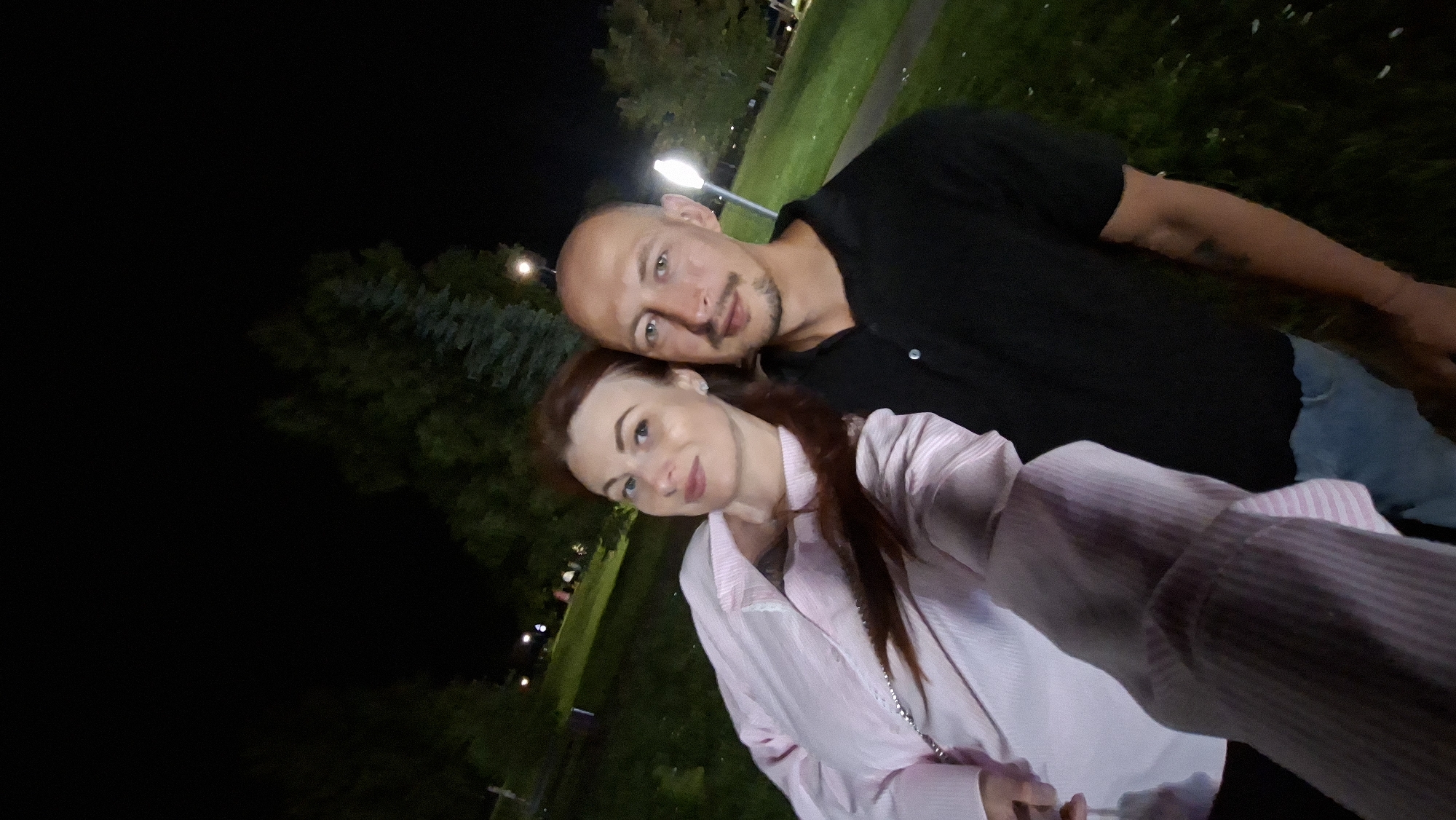 A selfie taken at night shows a man and a woman standing close together outdoors in what appears to be a park. The woman, on the left, has long reddish-brown hair and is wearing a light pink striped jacket. The man, on the right, is bald with a mustache and is wearing a black polo shirt. They are positioned in the foreground, with a grassy area and trees visible behind them, along with a lit lamppost. The environment suggests a public outdoor space in Myadzyel, Belarus, likely a park or recreational area, given the presence of manicured grass and trees, illuminated by artificial lighting.