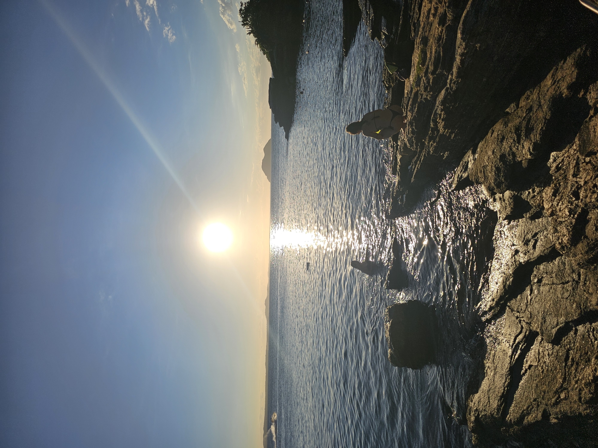 The image shows a coastal scene in Armação Dos Búzios, Brazil, at sunset. A woman is crouched on dark, rocky terrain at the water's edge, interacting with the ocean. The sun is setting over the water in the distance, creating a bright reflection on the water surface. There is also a person, presumed to be a swimmer, in the water. The background includes land, likely a shoreline or a distant landmass.