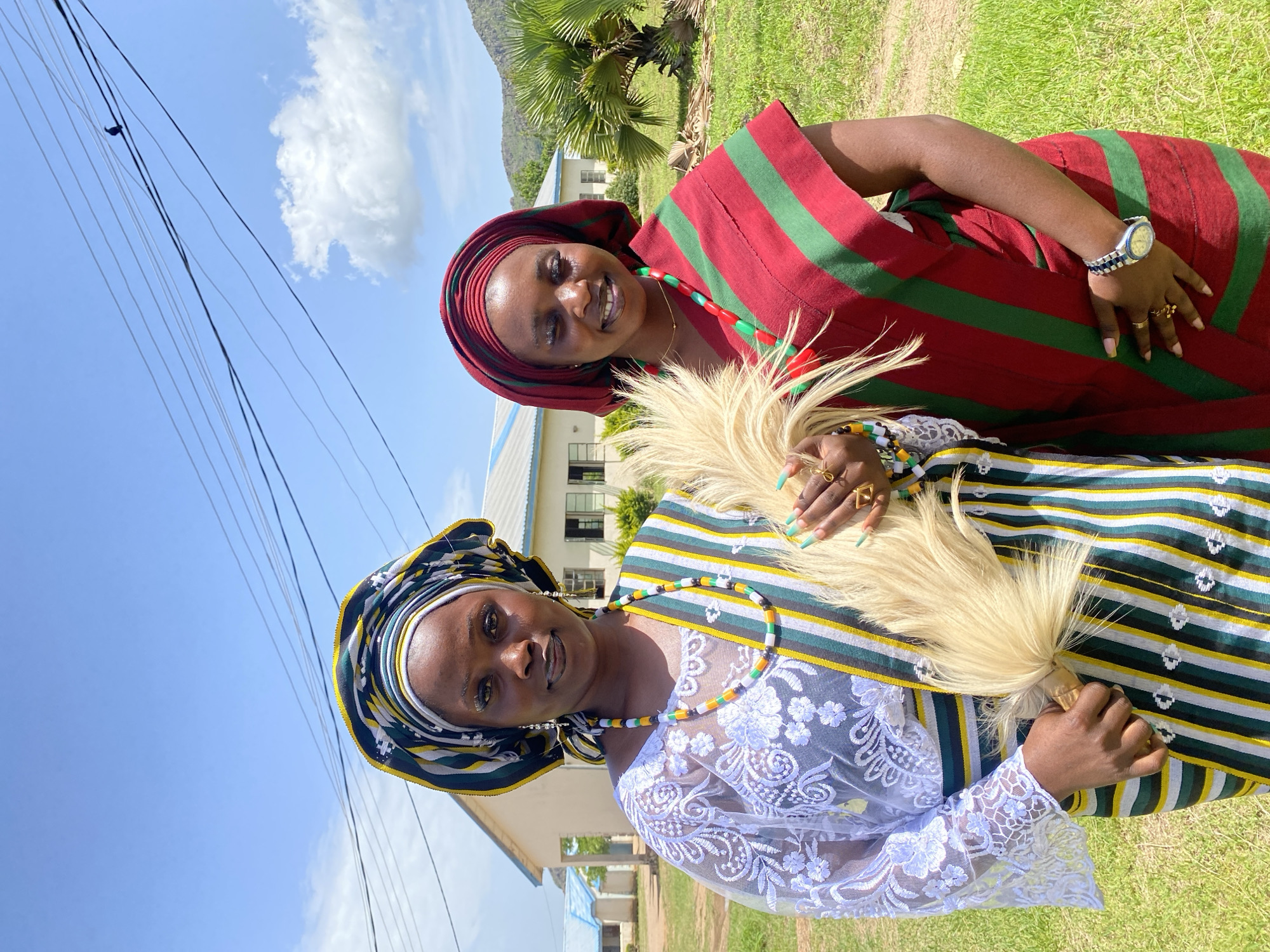 Two women stand outdoors, posing for a photograph. The woman on the left wears a white lace top and a striped headdress, a striped sash, and a necklace of colorful beads. She holds a light-colored, fan-like object. The woman on the right wears a red and green striped garment and a matching red headdress, and a red bead necklace. She is smiling. In the background, a light-colored building with blue trim is visible, along with green grass and trees. The sky is blue with scattered clouds. The location is identified as Wang, Nigeria.