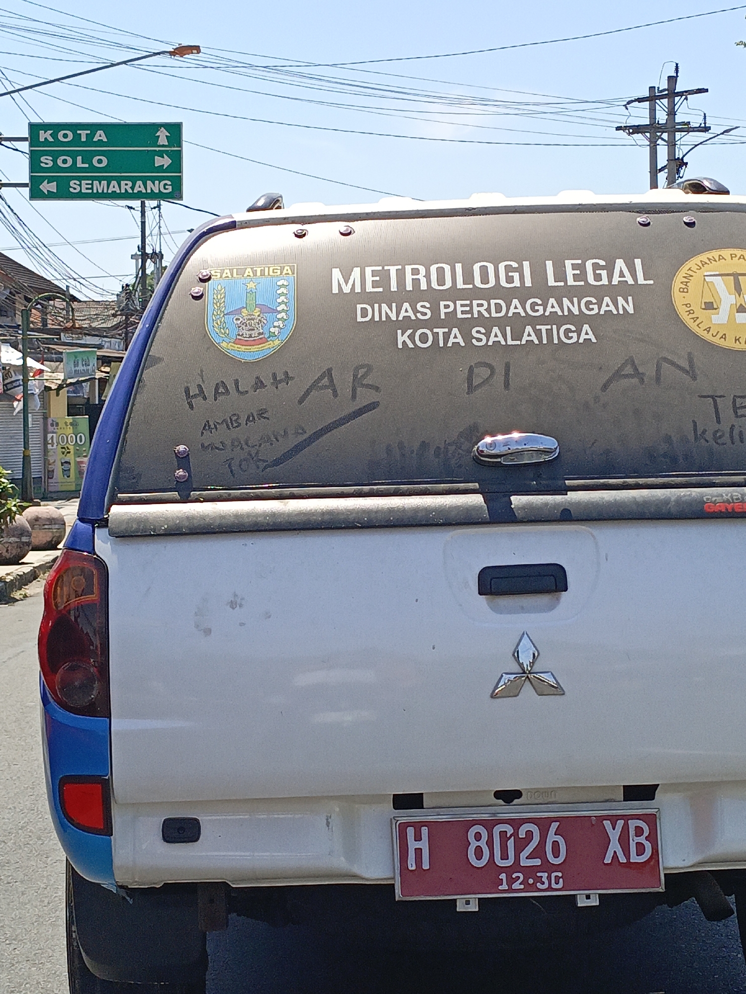 The image shows the rear of a white pickup truck in an outdoor urban environment. The truck has a dark-colored canopy with some graffiti scrawled on the rear window. A crest with the text "SALATIGA" is visible on the canopy. Text above the crest reads "METROLOGI LEGAL DINAS PERDAGANGAN KOTA SALATIGA." A red license plate with white lettering is mounted on the tailgate, reading "H 8026 XB" with "12.30" below.
To the left of the truck, a green street sign is mounted on a pole, indicating directions to "KOTA SOLO," "SEMARANG," and "KOTA." The background shows power lines and some buildings, suggesting a commercial or residential area. The ground appears to be paved. The overall lighting suggests it is daytime with a clear sky. The context of the truck's markings and the street sign indicate the location is within or near the city of Salatiga, Indonesia.