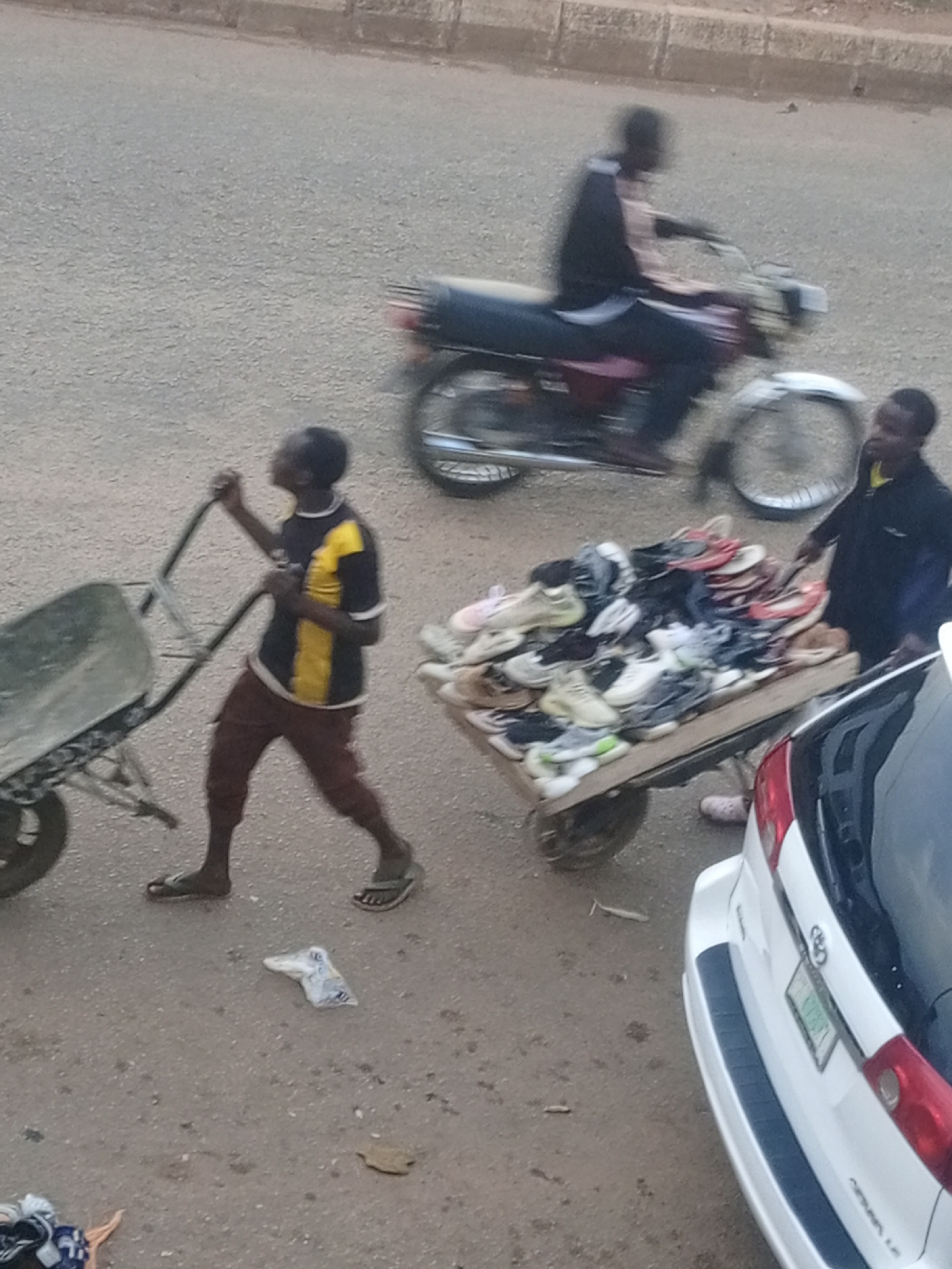 In Bukuru, Nigeria, a young man pushes a wheelbarrow filled with miscellaneous items, possibly construction debris, on a paved street. He wears a dark shirt with yellow and white accents and brown trousers. To his right, another young man stands by a cart overflowing with shoes of various styles and colors. Behind them, a motorcycle with a rider in motion passes by. In the foreground, the rear of a white SUV is partially visible. The scene depicts a busy street environment with elements of commerce and transportation.