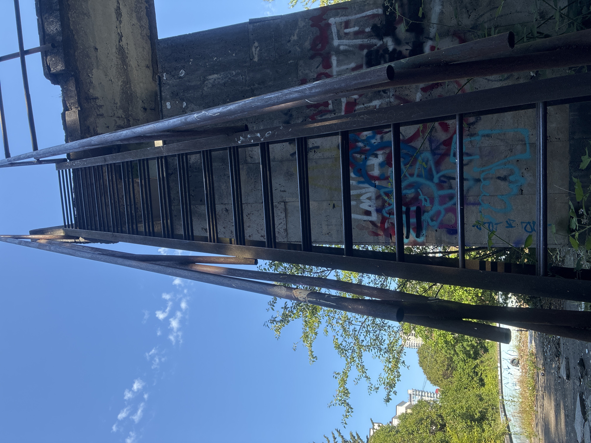 The image, taken in Sochi, Russia, displays an outdoor scene featuring a prominent metal structure resembling a steep ladder or grate, diagonally positioned against a backdrop of a clear blue sky and a graffiti-covered concrete wall. The metallic framework consists of dark, likely rusty, beams and numerous horizontal rungs or bars.
To the left and bottom of the frame, a vibrant blue sky with scattered white cumulus clouds suggests a bright, sunny day, likely mid-day. To the right and top, a weathered concrete wall is extensively covered with various graffiti tags in multiple colors, including red, blue, black, and white. While much of the graffiti is abstract, some discernible elements include stylized letters and shapes, such as a possible "LA V" in blue, and other indistinct words and patterns. Running above the main metal structure and parallel to the concrete wall are two dark metal pipes.
Below the concrete wall, dense green foliage, comprising trees and bushes, indicates an overgrown environment. Through the leaves, distant urban buildings are visible, confirming an urban or suburban setting. The scene evokes a sense of industrial or abandoned infrastructure, where natural elements and urban art converge. No human presence or activity is depicted, creating a static and somewhat desolate atmosphere.