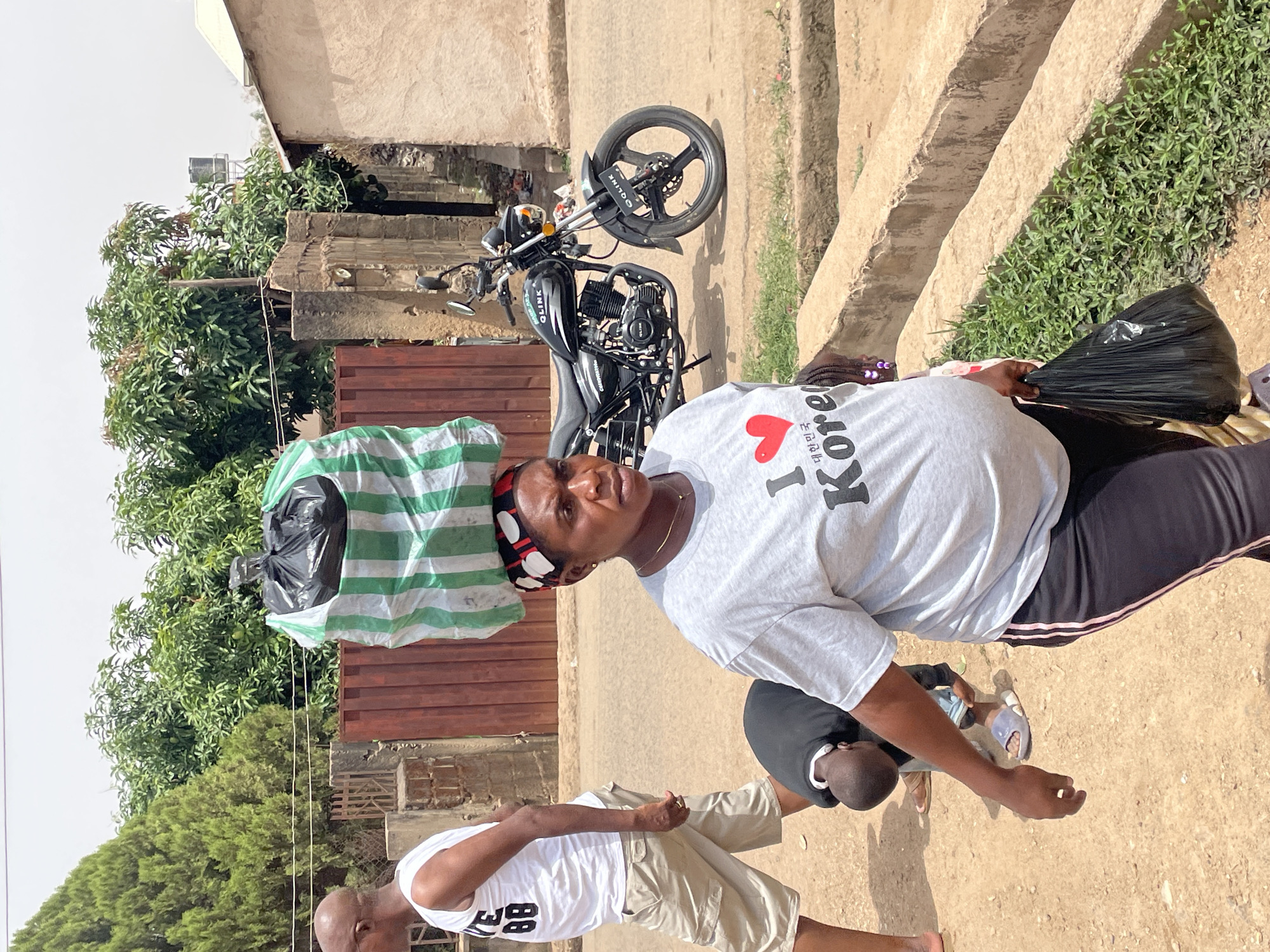 This sunlit outdoor image, taken in Jos, Nigeria, captures an everyday street scene with three people and a motorcycle. The photograph is oriented vertically, with subjects standing on a dirt path alongside a building and abundant greenery.
In the foreground, a woman faces the camera, wearing a light grey t-shirt that displays a red heart and the English text "I ❤️ Kore," followed by Korean characters (대한민국, meaning "Republic of Korea"). She has a patterned headwrap in red, black, and white, and dark trousers. She expertly balances a striped green and white bag, which contains a black plastic bag, on her head. In her right hand, she carries another black plastic bag. Her expression is neutral.
Behind her, a black Q-LINK motorcycle is parked, with its brand name visible on the fuel tank and front wheel fender. To the lower left, a man is partially visible, wearing a white t-shirt with the number "88" printed on it and khaki shorts, also looking towards the camera. Between the woman and the man, a third person, likely a child, is seen from behind, wearing a black t-shirt.
The background features concrete structures, including a building facade and what appear to be terraced concrete steps or an embankment on the right, covered in small green plants. Lush green trees, possibly mango trees, are prominent in the background, suggesting a residential or semi-urban environment. The bright sunlight and distinct shadows indicate it is midday or early afternoon with clear weather. The scene portrays a casual moment, possibly involving the woman transporting goods.