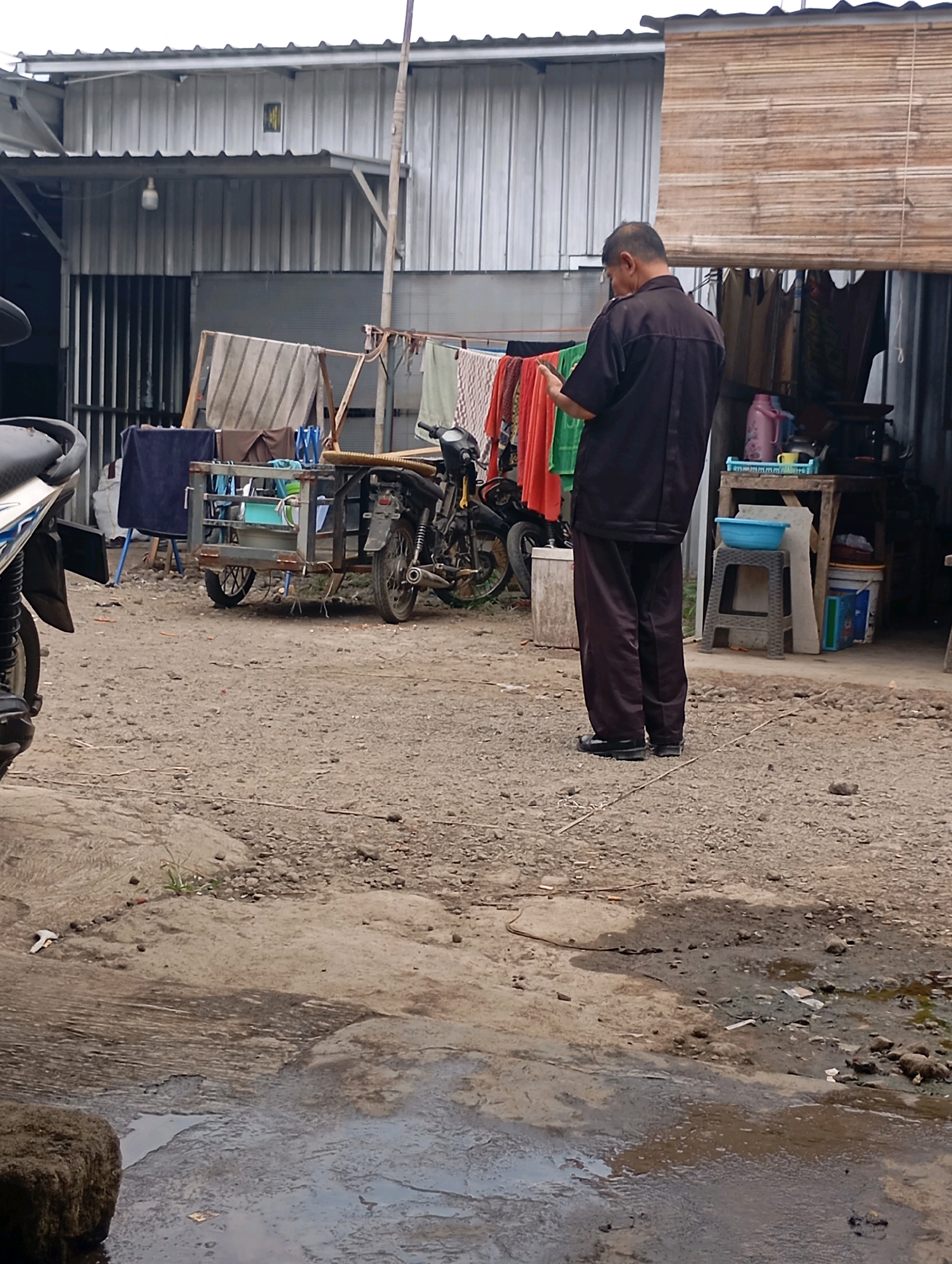 In an outdoor yard in Tasikmalaya, Indonesia, a man stands facing away from the camera, holding a smartphone. He is wearing a dark collared shirt and dark pants. The yard is unpaved and covered in gravel and dirt. To the left of the man is a motorcycle, and a small trailer with a blue tub is attached to it. Various items of clothing are hanging on a clothesline in the background, along with a bamboo blind covering a window. Further back, a building with a corrugated metal roof is visible. The ground in the foreground appears wet and muddy.