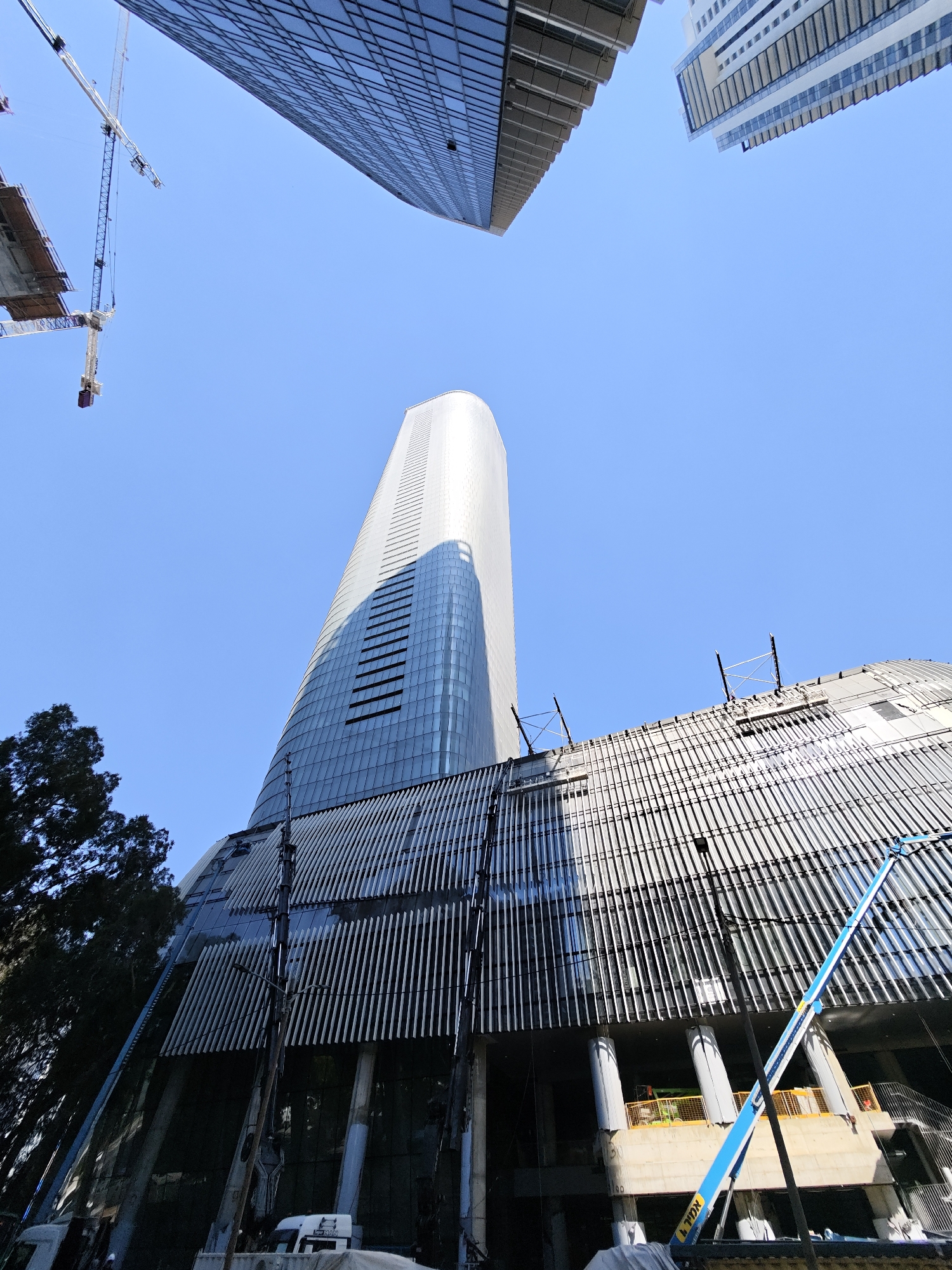 The image captures a construction site in Givatayim, Israel, dominated by the towering structures of modern skyscrapers against a clear blue sky. In the foreground, a distinctive curved building is under construction, its exterior featuring a pattern of vertical slats. A large blue boom lift is positioned near this building, its arm extended upwards. Two cranes are visible in the background, one partially obscured by a building and the other in the distance. In the upper portion of the frame, parts of two other skyscrapers are visible, one with a grid-like facade and the other with horizontal bands of windows. The scene suggests ongoing development in a bustling urban environment.