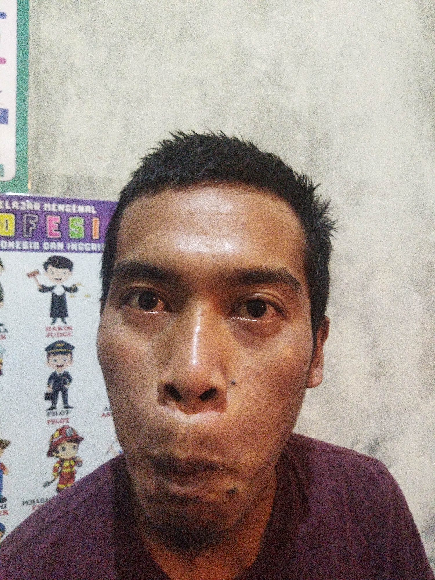 The image is a close-up, waist-up portrait of a man with dark, short hair, dark eyes, and a light brown complexion. He is making a fish-face, with his lips pursed inwards and his cheeks slightly puffed out. He is wearing a dark maroon t-shirt. The background appears to be a light-colored, textured wall, possibly concrete or plaster. To the left of the man's head, a portion of a poster with colorful graphics and text in Indonesian is visible. The text on the poster includes "BELAJAR MENGENAL" and "O FESI", and beneath it "ONESIA DAN INGGRIS". There are also small cartoon illustrations of various professions on the poster, such as a judge, a pilot, and a firefighter. The image is grainy, suggesting it was taken with a low-light setting or a lower quality camera. The lighting appears to be from the front, casting subtle shadows on the man's face. There are no discernible clues to indicate the specific location within Suci, Indonesia, beyond the presence of the Indonesian language poster.