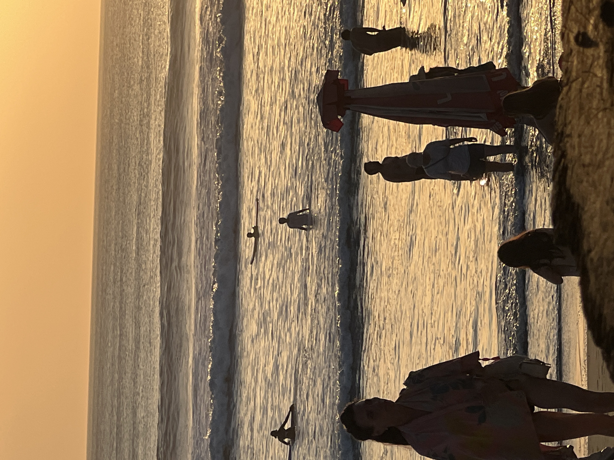 A wide shot captures a beach scene at sunset in Kuta, Indonesia. The sky is a soft orange, blending into the shimmering golden water. Gentle waves roll towards the shore, reflecting the warm light. Several people are visible in the water, some on surfboards and others wading. In the foreground, a woman in a patterned robe stands on the sand, facing the water. To her right, a beach umbrella, red and white, is partially obscuring a few individuals also standing in the shallow water. Silhouettes of people further out suggest they are surfing or preparing to surf. The overall atmosphere is serene and summery.