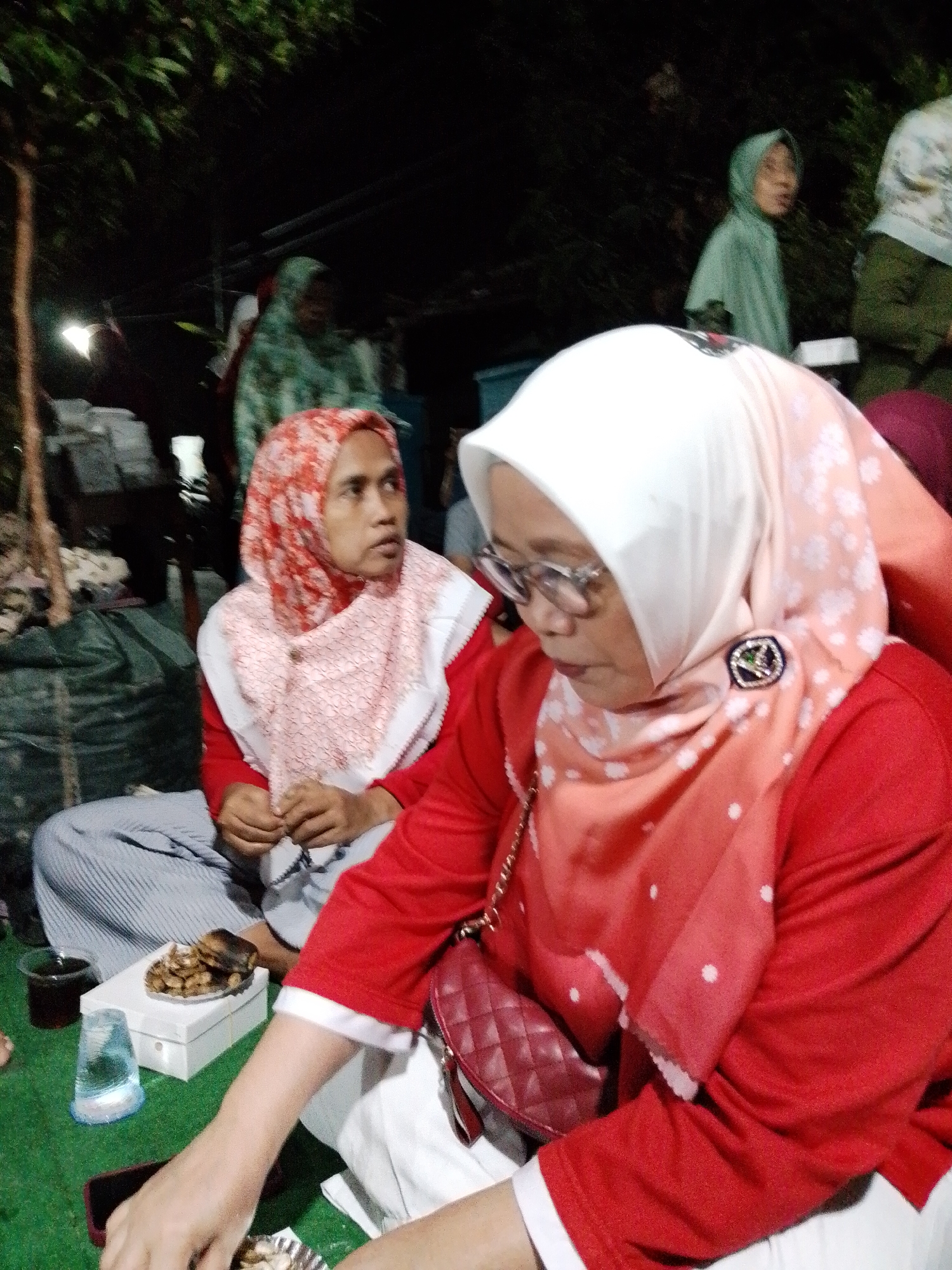 In Kecamatan Pati, Indonesia, at night, a group of people are gathered outdoors, likely for a community event or casual social gathering. The primary focus is on two women in the foreground wearing hijabs and traditional attire. The woman on the right wears a white hijab and a red top, with glasses and a decorative brooch. Her right hand is resting on a red quilted handbag. The woman on the left wears a red and white patterned hijab and a red top, and her hands are clasped in front of her, possibly holding prayer beads. Both women are seated on the ground, with a green matting covering the surface.
In the background, other individuals are visible, including one wearing a light green hijab and another in a green patterned hijab. There are also dark green bags and what appears to be some form of makeshift shelter or structure, suggesting an outdoor setting possibly in a less formal or public space. On the ground near the seated women, there is a white box, a clear plastic cup with a dark beverage, and a small plate of food items that appear to be fried snacks or crackers. The overall lighting suggests it is evening or night.