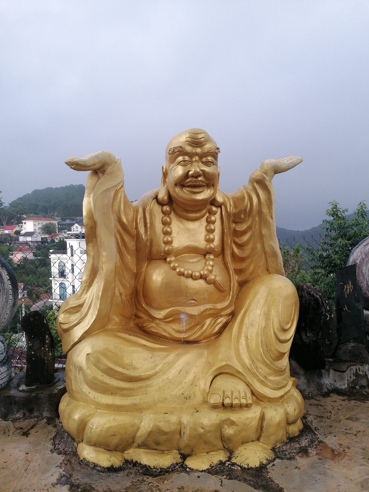 A large, golden statue of Budai, commonly known as the Laughing Buddha, dominates the foreground of the image. The Buddha is depicted with a wide, joyful smile, eyes squinted in mirth, and a prominent belly. He wears a flowing robe and a string of large, golden beads around his neck. His arms are raised and outstretched, with palms open in a welcoming or generous gesture, and he is seated in a relaxed, cross-legged posture. The statue rests on a textured concrete base, which shows some signs of wear and dark staining.
The setting is outdoors, overlooking a hillside town. In the mid-ground, numerous houses of various styles are visible, scattered across the verdant slopes, including a distinctive multi-story white building with arched windows. A chain-link fence runs horizontally across the scene, separating the immediate area around the statue from the distant residential structures. Beyond the houses, a range of lush, green hills and mountains recedes into the distance. The sky is uniformly overcast and grey, suggesting a cloudy day. The ground immediately around the statue is concrete. The scene conveys a sense of tranquility and joy, characteristic of a spiritual site in a scenic, elevated location like Da Lat, Vietnam. No discernible text is visible.