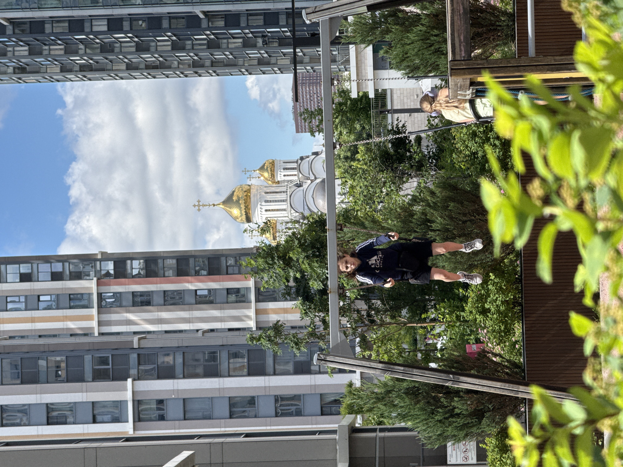 The image depicts a playground in Moscow, Russia, with a child on a swing in the foreground. Behind the playground, a large white building with golden domes, identifiable as a church, is visible against a blue sky with clouds. To the left and right of the church are tall, modern apartment buildings, characteristic of the urban landscape of Moscow. The child on the swing is wearing a dark blue hoodie and black shorts, with athletic shoes. Another person, possibly a girl, is visible on another swing in the background, wearing a light-colored shirt and shorts. Green trees and bushes surround the playground, creating a contrast with the urban structures. The overall scene captures a moment of recreation within a densely populated urban environment.
