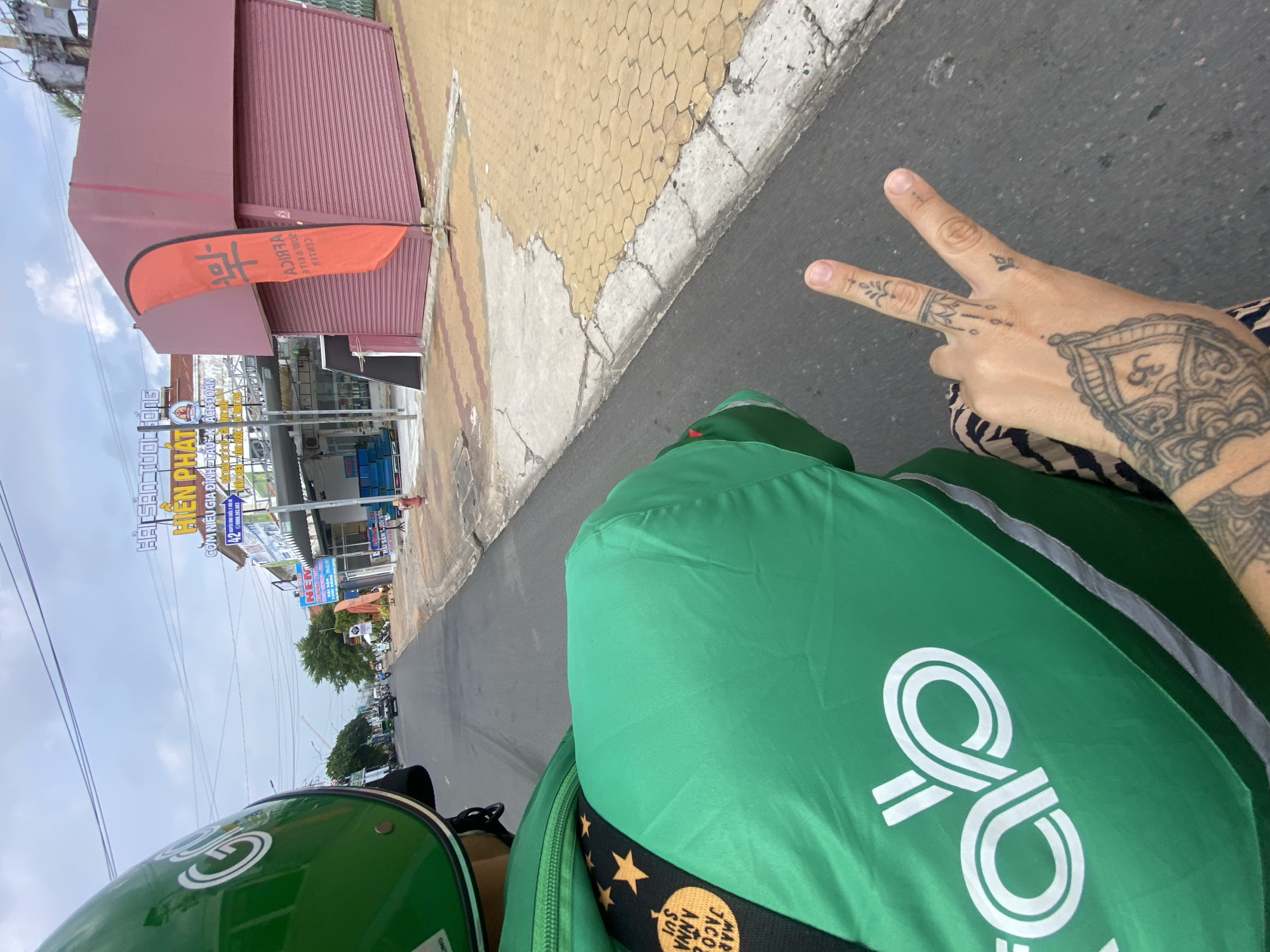 The image is a first-person perspective from the back of a moving motorcycle in Phu Thuy Ward, Vietnam. The rider is wearing a green helmet and a green Grab uniform with the Grab logo prominently displayed. A tattooed hand is visible, making a peace sign. The background shows a street with buildings, shops, and overhead power lines, typical of a Vietnamese urban environment. One building is painted maroon with a rolling metal door and an orange flag with black text, which appears to be in Vietnamese. Another building has signage that includes "HIEN PHAT" and "COM NIEU GIA DINH." The road surface is asphalt, and the sidewalk is paved with hexagonal yellow tiles. The sky is overcast.