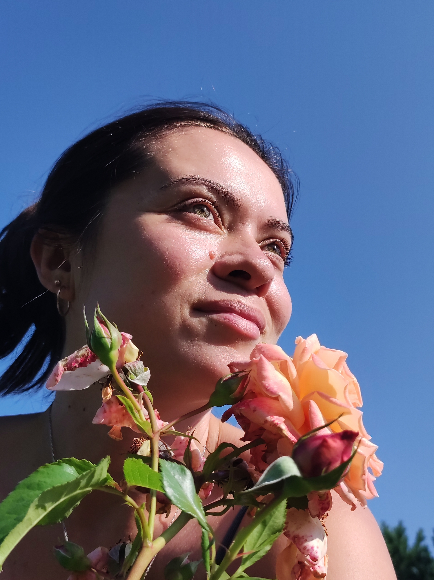 A young woman with dark hair and green eyes is positioned in the foreground, holding a cluster of pink and orange roses near her face. She is looking up and to the right, with a gentle smile. The sunlight illuminates her face, highlighting her features and casting a warm glow. The background is a clear, bright blue sky. There are also some green leaves and rose buds visible in the lower portion of the image. The overall setting appears to be outdoors, possibly in a garden. No specific location in the city of Фоки, Russia can be determined from the image.