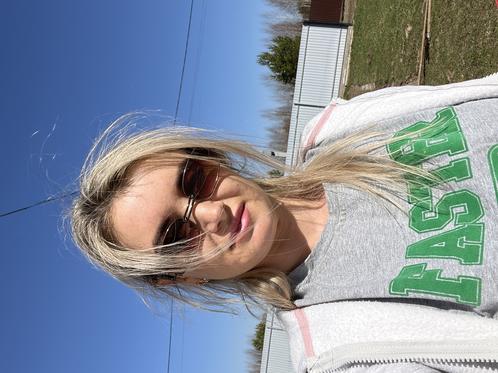 A woman poses for a selfie outdoors in Гаревая, Russia on a sunny day. She is wearing sunglasses, a grey top with green lettering that reads "FAST," and a white zip-up jacket with pink detailing. Her blonde hair is blowing in the wind. In the background, there is a fence, grass, a tree, and a clear blue sky. Power lines are also visible.