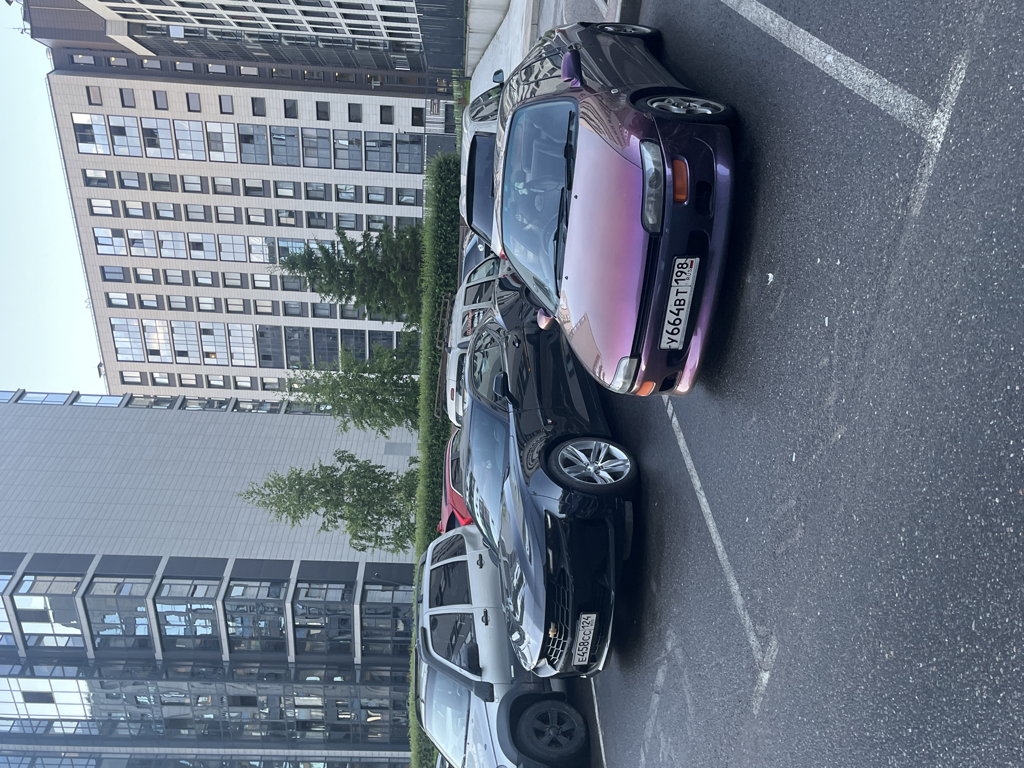 In the city of Unknown, Russia, two cars are parked on a wet asphalt surface in front of modern multi-story buildings. A black Chevrolet Camaro, license plate E458CC 124, is parked in the foreground. To its right, a purple Toyota Celica, license plate Y664BT 198, is also parked. A silver SUV is parked behind the Camaro. The buildings in the background are tall with numerous windows and balconies, reflecting the sky. Trees are visible between the buildings and the cars.