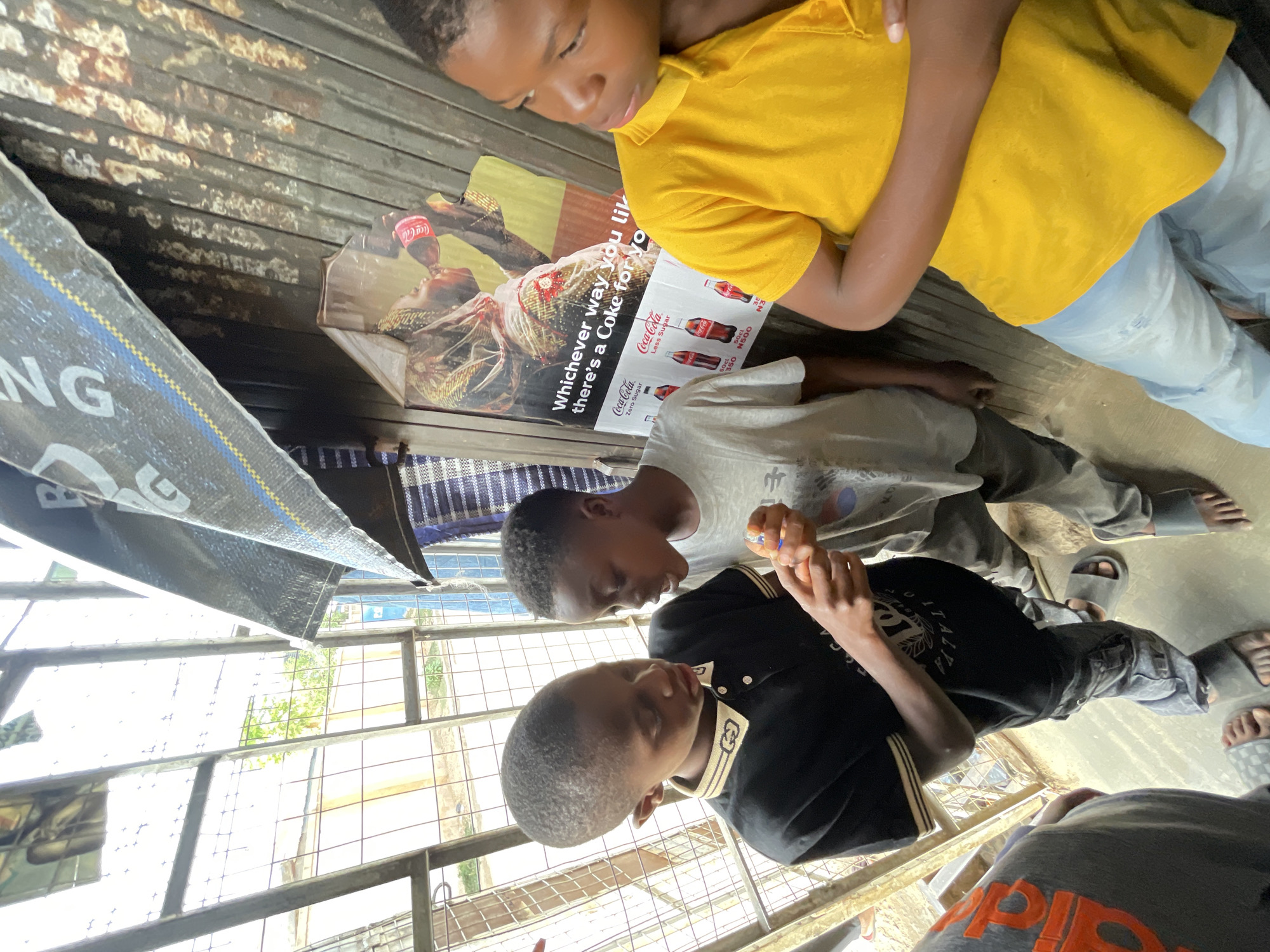 The image depicts three young boys in an outdoor setting. One boy wears a yellow shirt and light jeans, while another is in a black shirt. The third boy wears a grey shirt. Two of the boys are looking down at something in their hands. They are positioned near a Coca-Cola advertisement that includes the tagline, "Whichever way you like it, there's a Coke for you." The area has a metallic, corrugated surface with what appears to be a tarpaulin overhead. The location is suggested to be Jos, Nigeria. A window with a view of a building and trees is visible in the background.