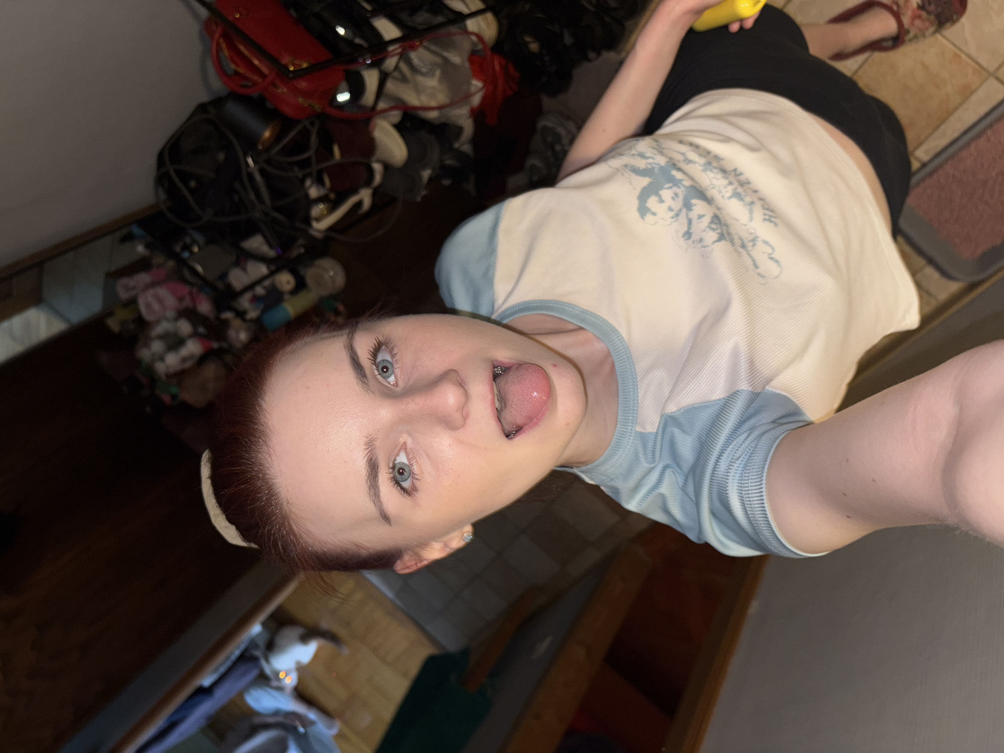 The image features a young person with red hair, blue eyes, and braces, looking directly at the camera with their tongue sticking out. They are wearing a light-colored t-shirt with blue accents and a graphic print. The person is holding a bright yellow object, possibly a toy or a piece of fruit, in their right hand. The background is cluttered with various items, including bags, shoes, and other personal belongings, suggesting an indoor setting, likely a home. Some of the items appear to be in disarray, indicating a lived-in space. The lighting in the image is dim, suggesting it might be evening or night. The overall tone of the image is playful and informal, with the person's expression conveying a sense of silliness or a lighthearted mood. The image orientation is rotated, with the person appearing to be lying down or viewed from a high angle. There is no visible text in the image.