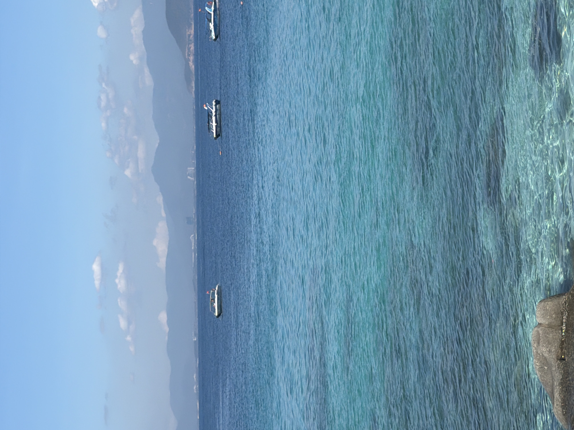 The image depicts a coastal scene in Nha Trang Ward, Vietnam, with a view of the ocean and the sky. Three boats are visible on the water. The water appears calm and clear, transitioning from a darker blue further out to a lighter, more turquoise color closer to the coast. A portion of a rocky shoreline is visible on the right side of the frame. In the background, a hazy skyline and mountains are visible. The sky is blue with some scattered clouds.