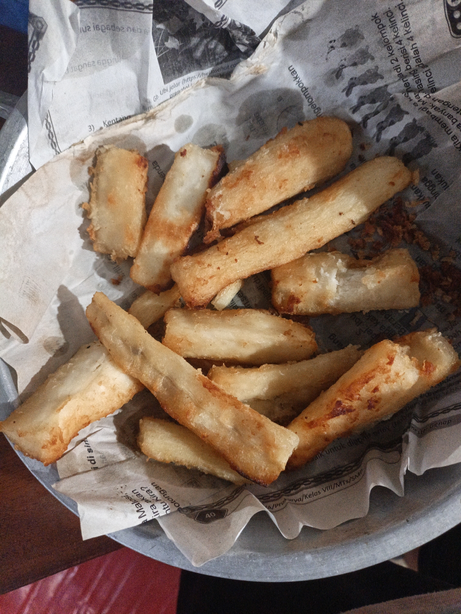 A close-up overhead shot shows a pile of fried cassava sticks, or "singkong goreng," arranged on a metal plate lined with newspaper. The cassava sticks are golden brown and appear to be lightly coated in a thin batter, suggesting they were deep-fried. Some pieces have visible specks of seasoning. The newspaper lining the plate is a common way to serve this snack in Indonesia, often used as a simple, absorbent base. The text on the newspaper is in Indonesian and appears to be from an educational or informational publication, with some words like "Kentang" (potato), "Kelompok" (group), and "Kelas" (class) visible. The background is out of focus but suggests an indoor setting with a wooden table and a hint of red fabric. The image captures a casual presentation of a popular Indonesian street food. While the specific location in Pare, Indonesia, is not definitively identified by visual cues in the image itself, "singkong goreng" is a widely available snack throughout Indonesia, including in cities like Pare, which is known for its educational institutions and associated student culture where such snacks are commonly consumed.