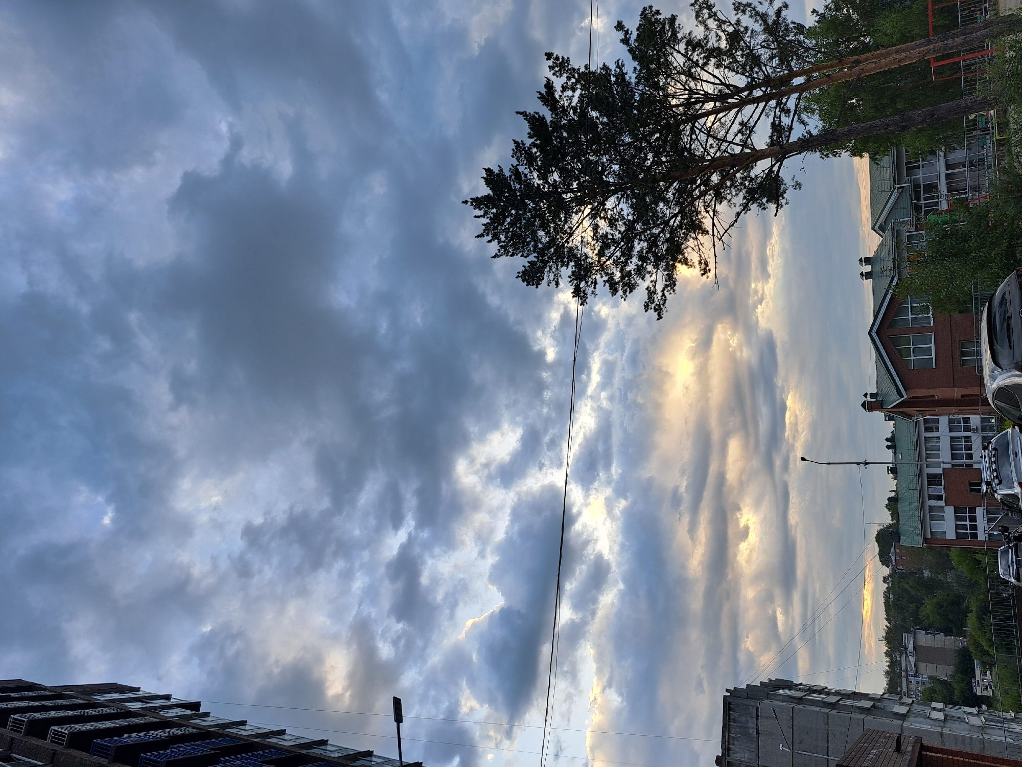 The image depicts a cloudy sky over an urban landscape, possibly in Irkutsk, Russia, as suggested by the prompt. In the foreground on the left, a tall, modern apartment building with balconies is visible. To the right, two tall pine trees are silhouetted against the sky. In the midground, several residential buildings with green roofs are present, along with some trees and a lamppost. The sky is filled with dramatic, dark gray clouds with patches of lighter, brighter clouds where sunlight breaks through, indicating it might be late afternoon or early evening. The overall atmosphere is somewhat somber due to the cloudy conditions. There are no people or discernible actions visible in the image.