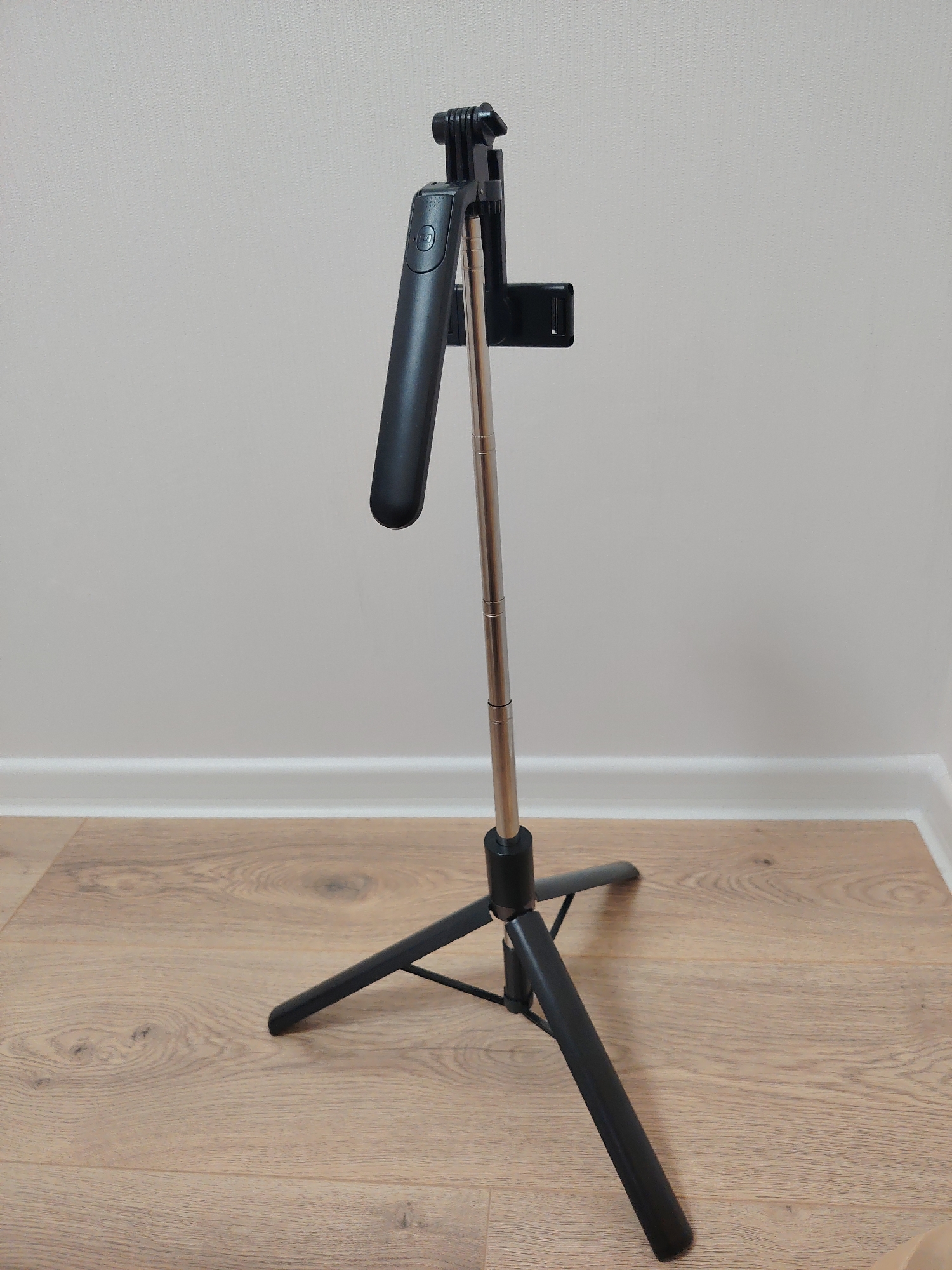 A single black and metallic gray selfie stick, configured as a tripod, stands upright on a light-colored wooden laminate floor. The device's metallic telescoping pole is partially extended, topped by a black smartphone mount. A detachable black oval-shaped remote control is docked into the handle section of the selfie stick. The three black legs of the integrated tripod are splayed outwards for stability on the floor. The floor features a visible wood grain pattern and meets a clean white baseboard. Behind the device is a plain, light grey wall with a subtle, uniform texture. The scene is brightly and evenly lit. No individuals or other distinguishing objects are present. The specific location within the city of Yekaterinburg, Russia, cannot be determined from the visual information provided.
