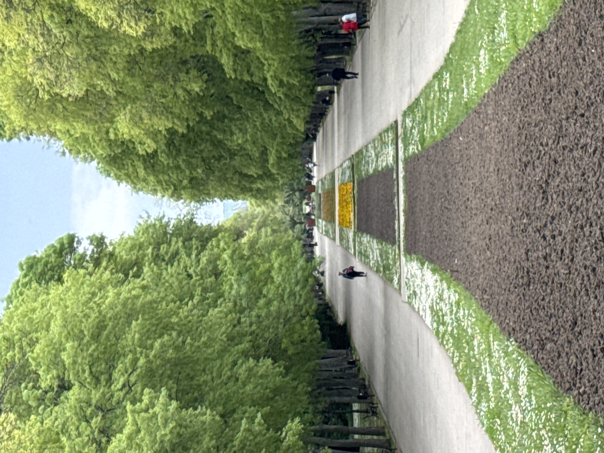The image shows a long, straight pedestrian walkway lined with trees, likely located in a park in the city of Varna, Bulgaria. The walkway is flanked by rectangular flowerbeds, some containing flowers and others appearing to have dark soil. People are walking along the walkway. Benches line the sides of the walkway between the flowerbeds and the trees. The sky is visible in the distance between the tree canopy. The photo is taken at ground level with a shallow depth of field.