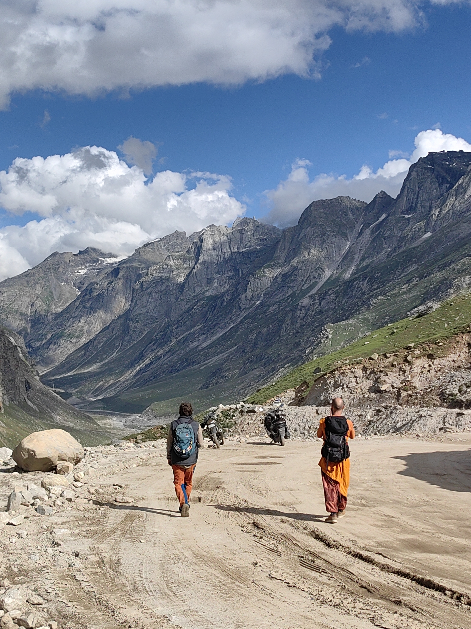 In a mountainous landscape, two individuals walk away from the viewer on a dirt road. The person on the left, wearing a backpack and orange pants, walks ahead. The person on the right, dressed in an orange robe and also wearing a backpack, follows closely. Two motorcycles are parked on the road behind them. In the background, steep, rocky mountains rise up, with green vegetation on some slopes. The sky above is blue with scattered white clouds. The location is specified as the city of Bashisht, India.
