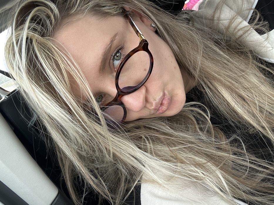 An adult female is depicted in a close-up, self-portrait style photograph, taken within what appears to be a vehicle in Torbalı, Turkey. The subject, possessing long, multi-toned blonde and light brown hair, is positioned with her head slightly tilted, and her hair is somewhat disheveled or windblown, obscuring a portion of her forehead and left side of her face. She wears oval-shaped eyeglasses with tortoise-shell frames, through which her light-colored (likely green) eyes are visible, maintaining direct eye contact with the camera. Her lips are pursed in an expressive manner. A small, round, silver-colored stud earring is present in her visible right earlobe. Her attire includes a dark garment layered under a lighter-colored, possibly beige or off-white, outer garment which could be a hoodie or jacket. The visible background suggests the interior of a vehicle, with a white or light-colored panel or trim element visible on the upper left, and darker, indistinct shapes behind her head.