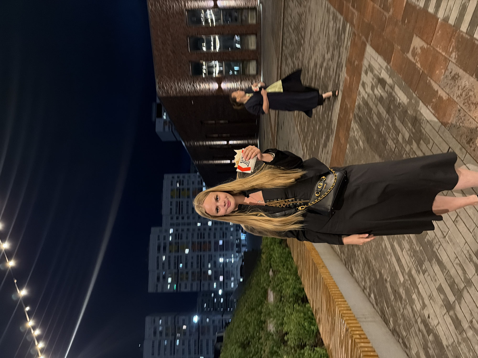 A young woman with long blonde hair and a black dress stands on a paved walkway in a city setting at night. She is holding a small box with a red and white logo. Behind her, a woman in a dark blue dress walks away with a cup in her hand. In the background, there are modern apartment buildings and a brick building with illuminated windows. String lights are visible overhead. The ground is wet, suggesting recent rain. The location is Pervomaysky, Russia.