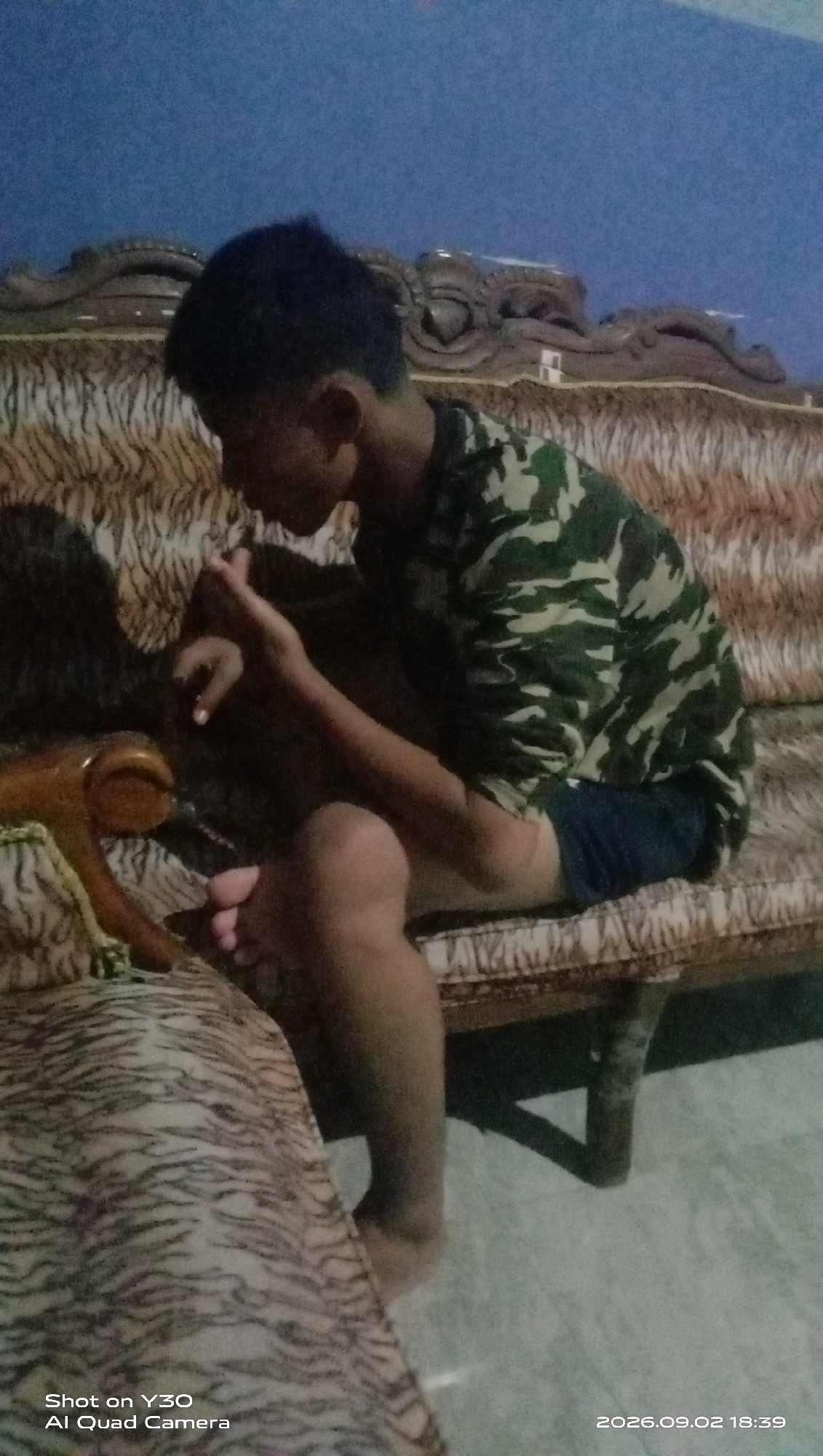 A young male is sitting on a patterned sofa in a room. The sofa has a tiger-stripe pattern. The male is wearing a camouflage shirt and dark shorts. He is looking down and appears to be holding something in his hands. The background is a dark blue wall with some decorative carvings on the top. The lighting in the room is dim. A timestamp in the bottom right corner of the image indicates the date is September 2, 2026, and the time is 18:39.