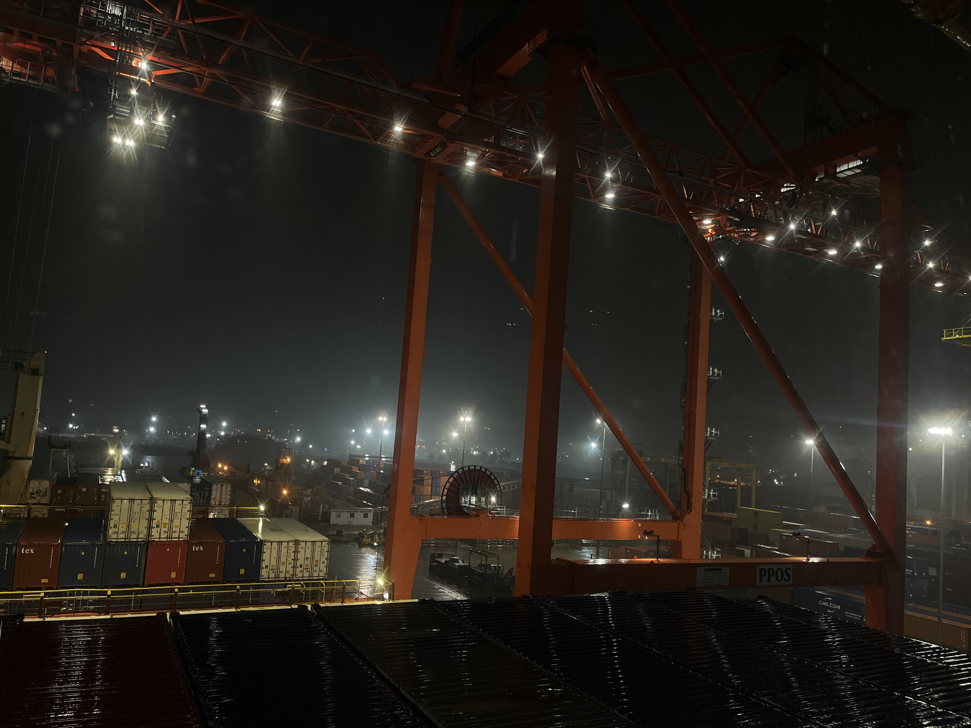 A low-angle, wide shot captures a container port at night. The foreground is dominated by the dark, wet, ridged surface of stacked containers. Rising above, a large, orange container crane stands prominently, its structure illuminated by numerous lights. In the background, containers are stacked in rows. Streetlights dot the area, reflecting off the wet pavement. The overall environment is dark and industrial, with the focus on the logistics and infrastructure of a busy port. No people are visible. The city is unknown.