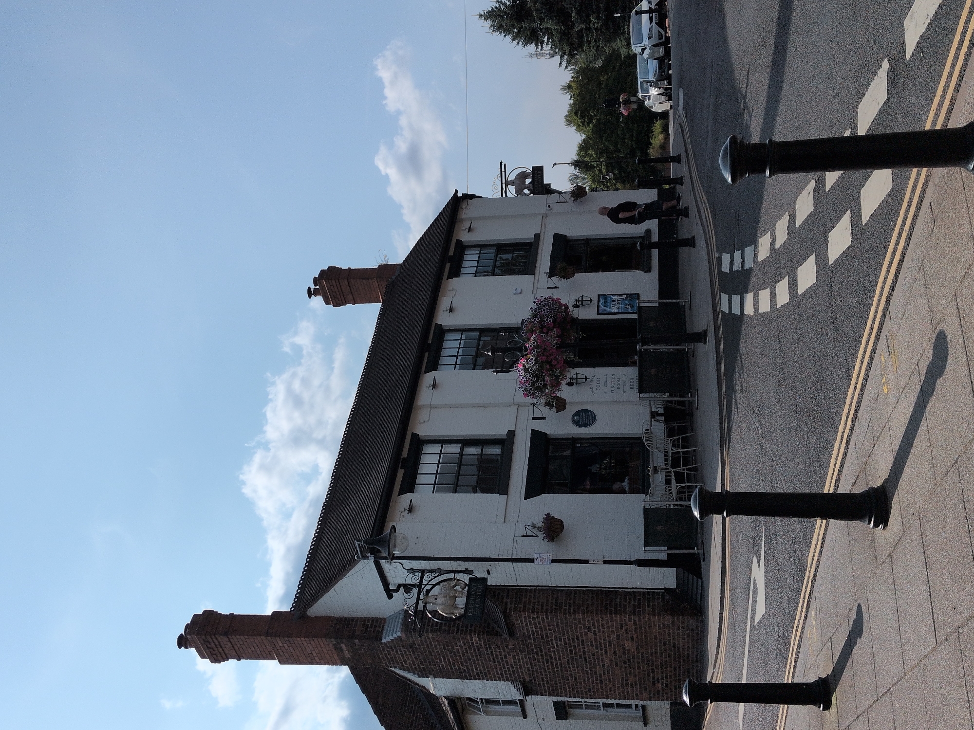 The image depicts a white building with a dark, pitched roof, situated alongside a paved street. The building features several windows, some with dark frames, and a brick chimney. Hanging baskets with pink flowers are visible on the exterior. A sign with a white circular emblem and text hangs from the building. Several black bollards line the edge of the street, which has a yellow double line marking. White diamond shapes are painted on the asphalt. In the background, trees and parked cars are visible. The sky is blue with scattered white clouds. The sun appears to be casting long shadows, suggesting it is either early morning or late afternoon. The scene is quiet and appears to be a street in a town or village. There are no people clearly engaged in any activity. Visible text includes "FUNCTION HIRE FOOD BEER" on a sign at the top of the building.