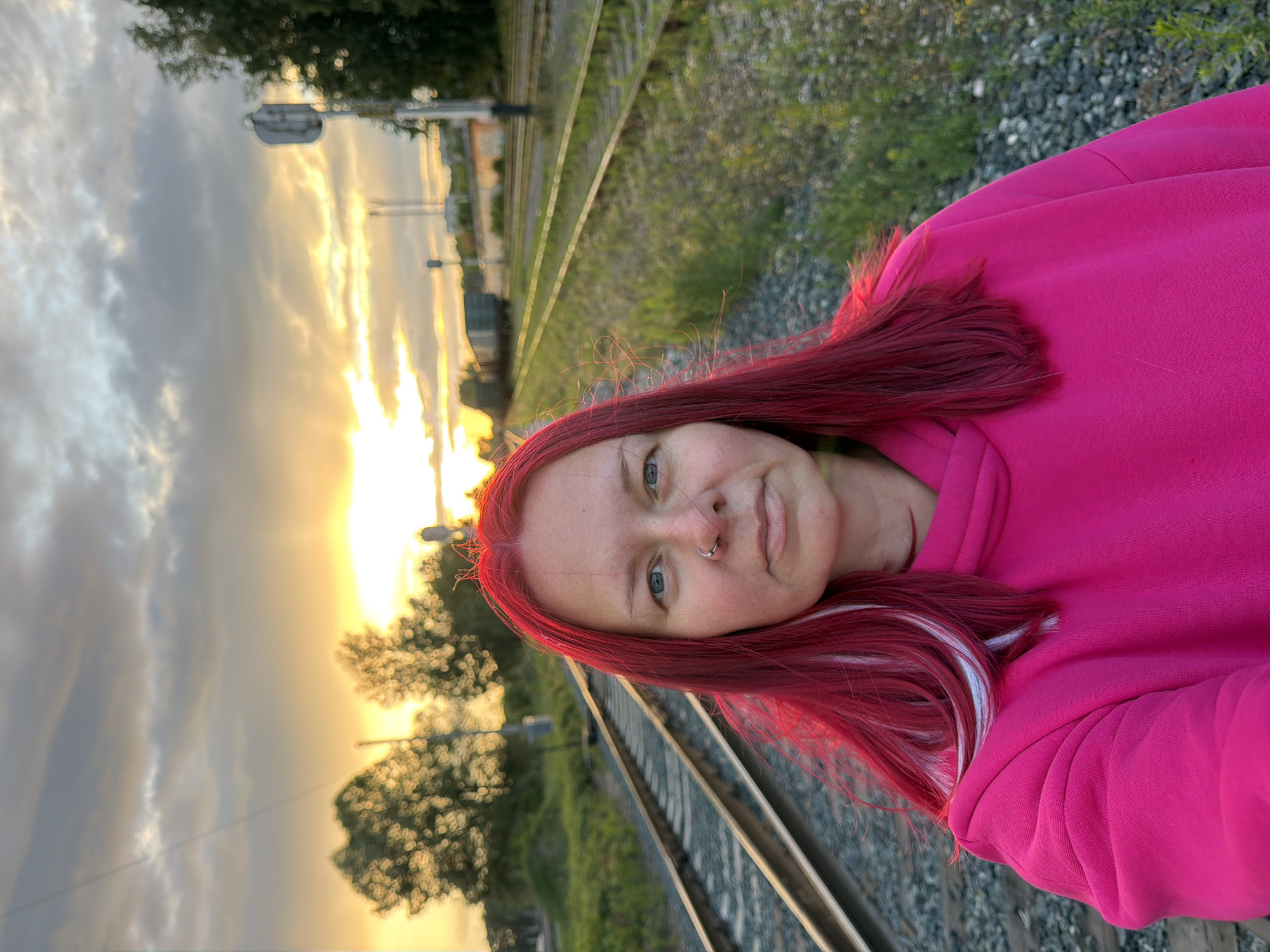 A selfie is taken from a low angle, showing a person with bright red hair and a nose ring. They are wearing a vibrant pink hooded sweatshirt. The background features train tracks running diagonally across the frame, with lush green vegetation on the right. The sky on the left is filled with dramatic clouds illuminated by a bright, setting sun, casting a warm, golden glow. The overall scene suggests an outdoor setting at dusk. The person's expression is relaxed and neutral.