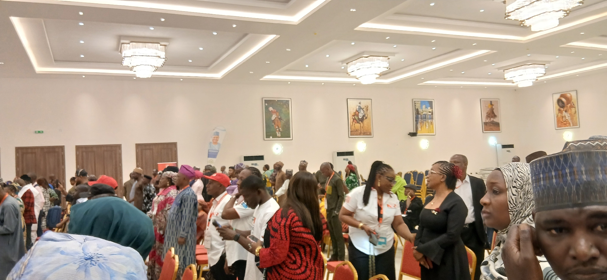 A wide-angle shot taken indoors shows a large group of people assembled in a well-lit room with high ceilings. The walls are painted in a light color and adorned with several framed pictures spaced apart, each featuring different subject matter. Near the back, three sets of double doors are visible. The room is illuminated by multiple chandeliers with a modern design. The people are diverse in age and dress. Some are standing, while others appear to be seated. Their actions and interactions are varied, with some conversing, some looking at their phones, and some seemingly observing the scene. The location appears to be a large hall or event space.