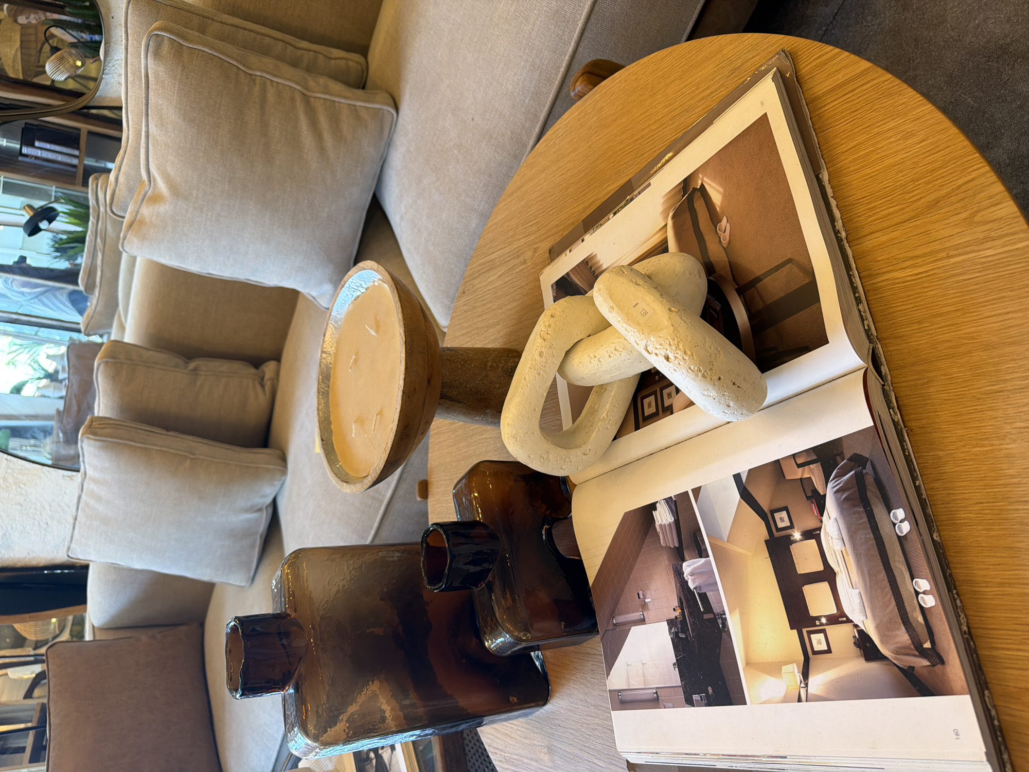 The image displays a cozy living room scene featuring a plush, neutral-toned sofa adorned with several throw pillows. In the foreground, a round wooden coffee table holds a collection of decorative items. Two amber glass vases, one larger and rectangular, and the other smaller and more uniquely shaped, are placed on the table. Next to them, a wooden bowl filled with a pale, creamy substance, possibly a candle or potpourri, rests on a wooden pedestal. A stack of what appears to be a hardcover book or magazine is open on the table, revealing interior design photographs of a bedroom and bathroom. A white, knotted decorative object made of a stone-like material is positioned atop the open book. The overall ambiance suggests a modern and stylish interior design. The image does not provide any specific landmarks or context that would indicate a location in Tel-Aviv, Israel.