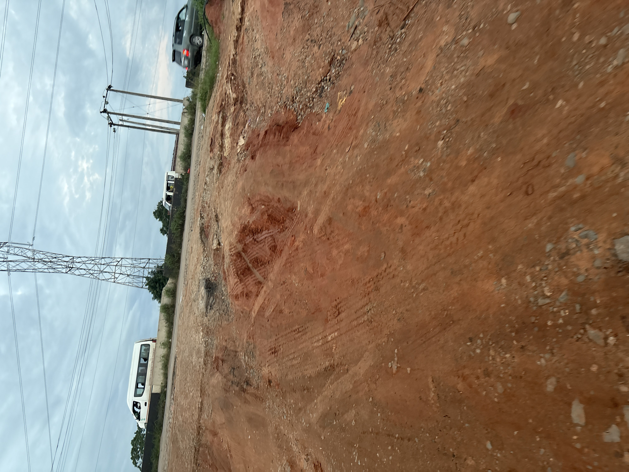 The image captures a cloudy day scene in Orifite, Nigeria, with a focus on a construction or excavation site in the foreground. Reddish-brown soil dominates the lower half of the frame, showing evidence of tire tracks and uneven terrain, suggesting recent vehicular activity and earthmoving. In the background, a white minivan with tinted windows is visible on a raised road, indicating a transportation route. Further back, a utility pole with multiple wires stretches across the sky, alongside another, larger electricity pylon. A dark-colored SUV is also parked on the roadside in the mid-ground. The sky is overcast with visible clouds, contributing to the subdued lighting of the scene. The overall impression is of an active construction zone on the outskirts of a town, with infrastructure and vehicles indicating movement and development.