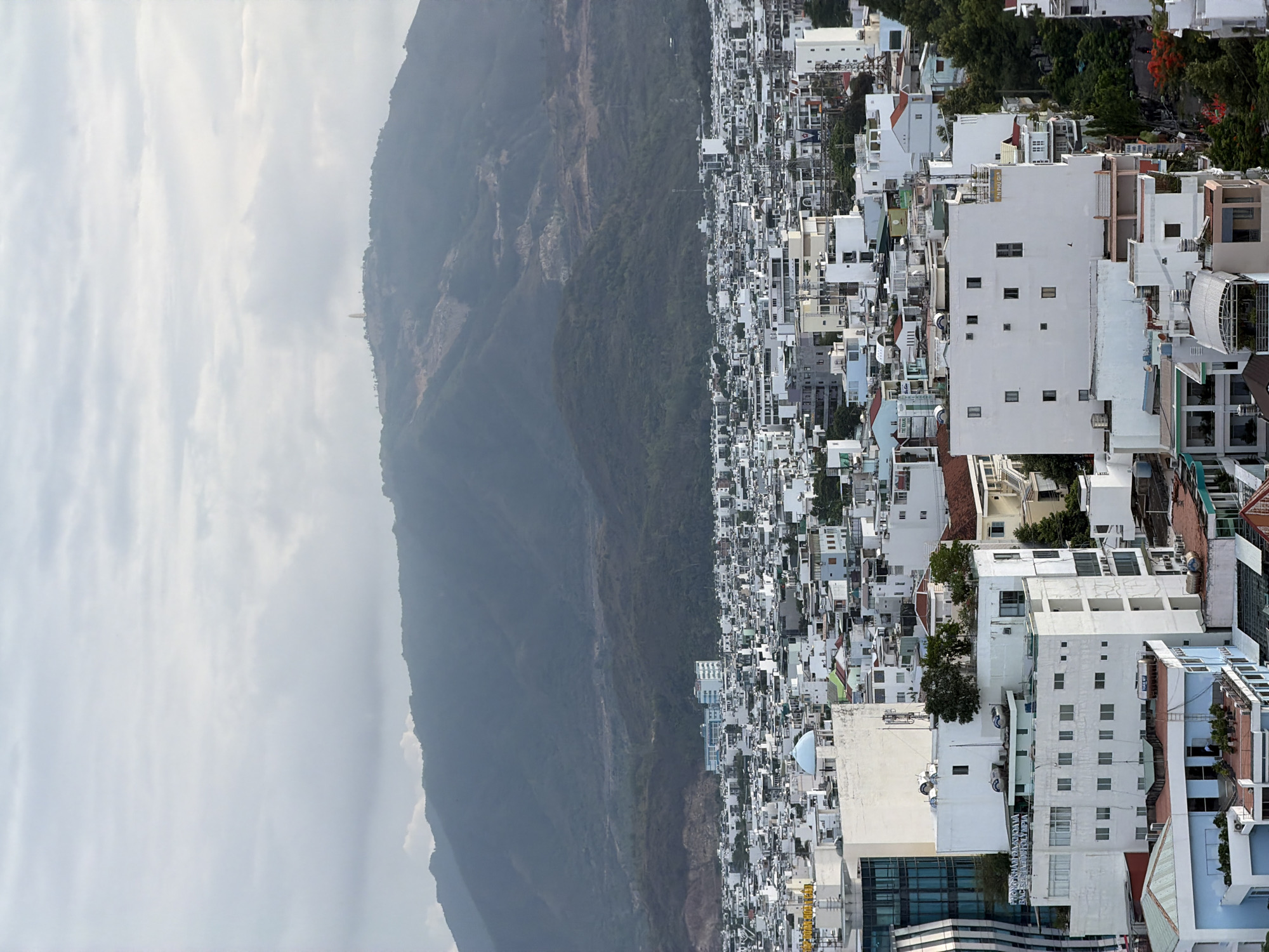 The image presents a panoramic, high-angle view of a densely developed urban landscape situated at the base of a towering mountain range under an overcast sky. The scene depicts a city, specifically identified as Nha Trang Ward, Vietnam, extending horizontally across the frame.
The city is characterized by a multitude of buildings, primarily multi-story structures painted in white or light neutral tones, with various architectural styles that appear modern and functional. These buildings are tightly packed, forming a dense urban fabric with visible streets and pathways winding through them. Green trees and other vegetation are interspersed among the buildings, adding pockets of color to the predominantly white cityscape. Some buildings feature balconies, varied roof designs, and occasionally blue-domed structures.
Dominating the background is a vast, dark green mountain range, its slopes covered in dense vegetation and hints of exposed rock. The mountains rise steeply, merging with the cloudy, uniformly gray sky which indicates an overcast daytime. A slight haze or atmospheric perspective softens the distant mountain peaks.
Visible text on several buildings confirms the Vietnamese location. On one white building, the characters "AN PHÚC CÔNG" are partially legible. Another large building features a prominent blue sign with white Vietnamese text, which appears to read "NGÂN HÀNG" (Bank) and "HỘI SỞ GIAO DỊCH" (Transaction Office), suggesting a bank branch.
No specific human activity or event is discernible, providing a static yet detailed snapshot of this bustling Vietnamese city nestled between mountains and, implicitly, the coast.