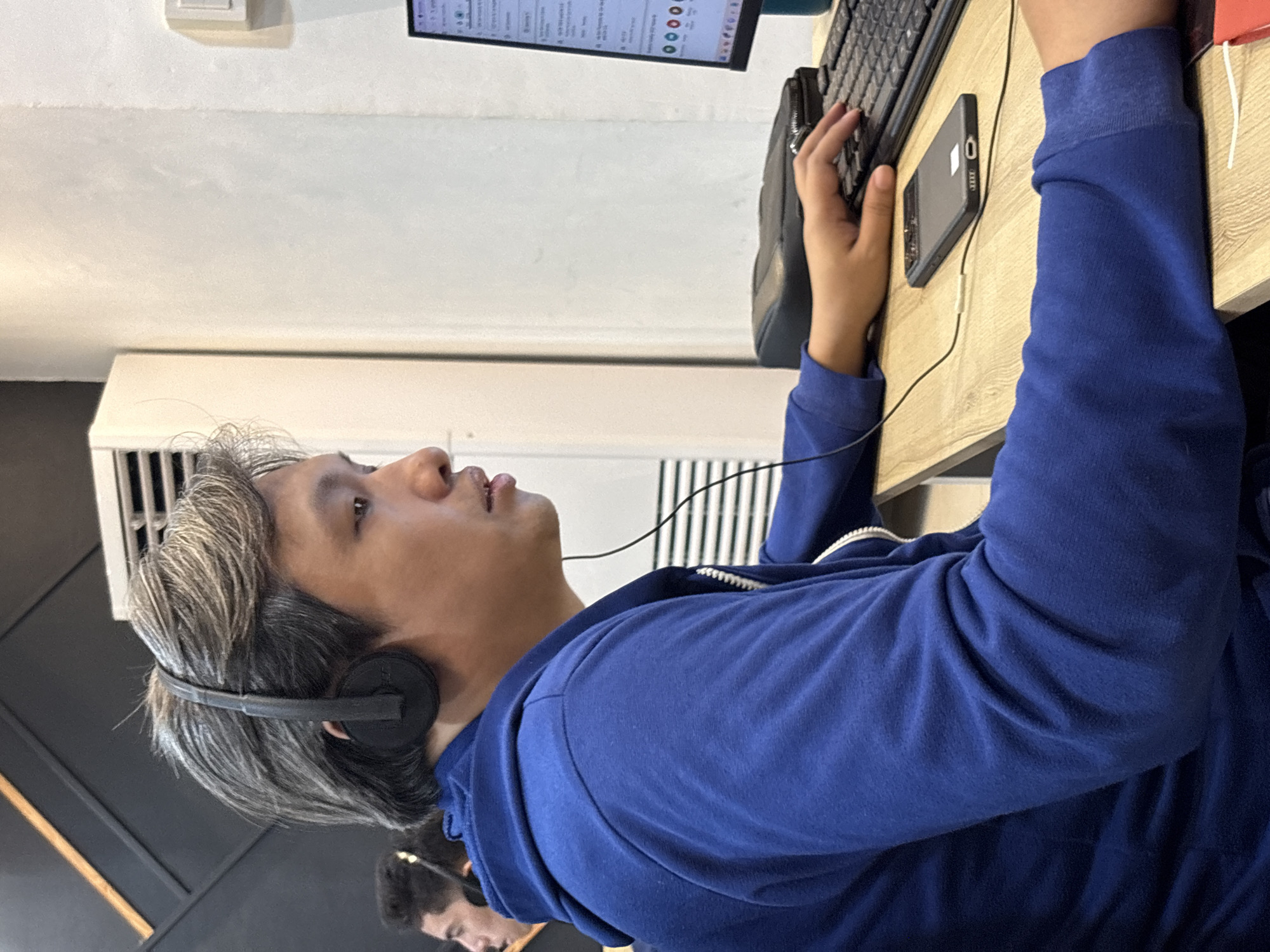 In Davao City, Philippines, a person with graying hair, wearing headphones and a blue hooded jacket, is seated at a wooden desk. They are looking towards a computer monitor, with their right hand positioned over a keyboard, seemingly typing. A smartphone is placed on the desk in front of them, connected by a wire to a power source or device. The background shows a wall with a white air conditioning unit and some dark geometric patterns. The environment suggests an indoor workspace.