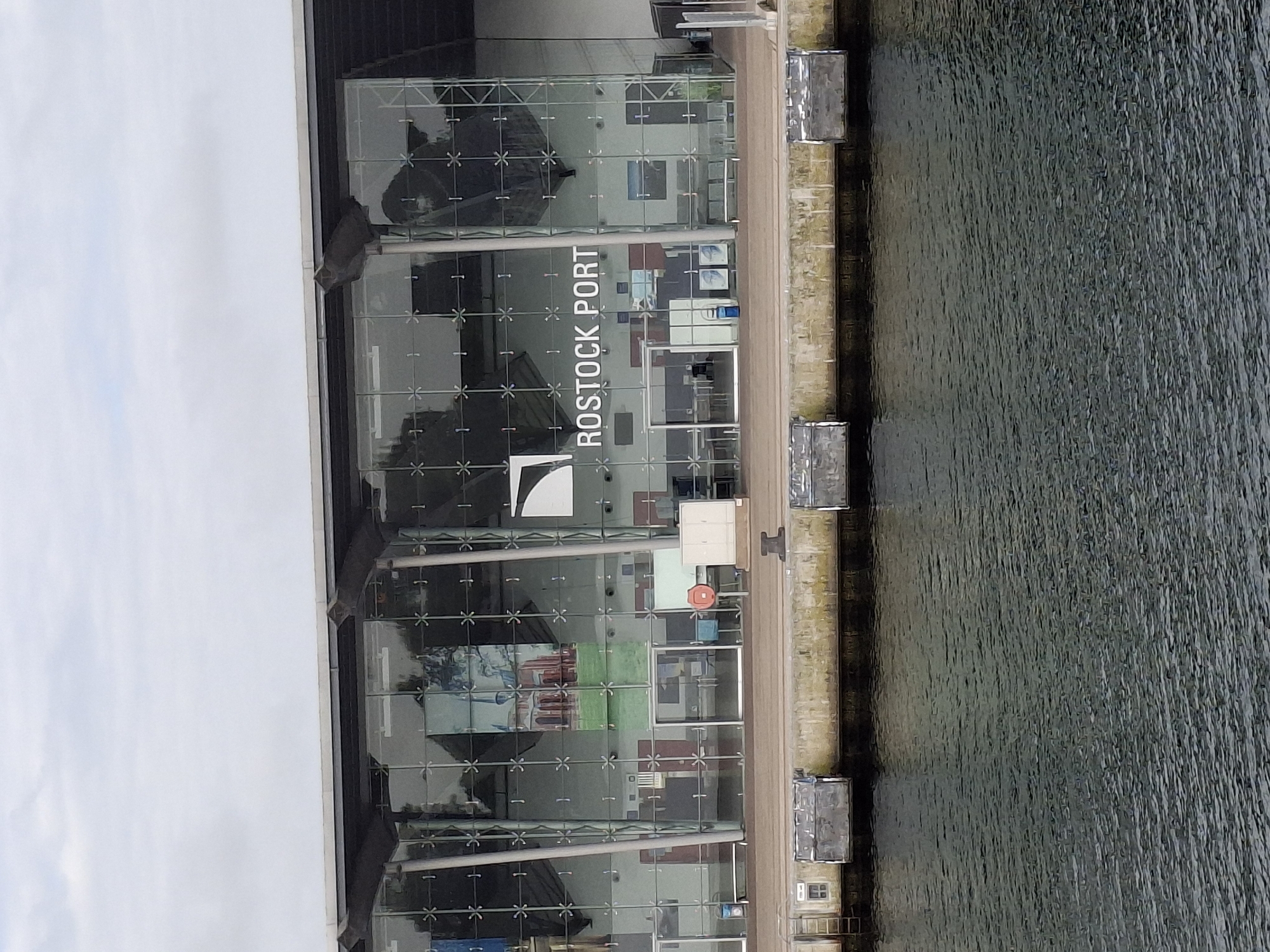 A sideways aerial-like view captures the Rostock Port building in Germany, situated beside water. The building is modern, featuring a glass facade divided into a grid pattern. The words "ROSTOCK PORT" are written vertically on the exterior of the building. A walkway runs along the side of the building adjacent to the water. The water is dark and has a slightly rippled texture. The sky is overcast.