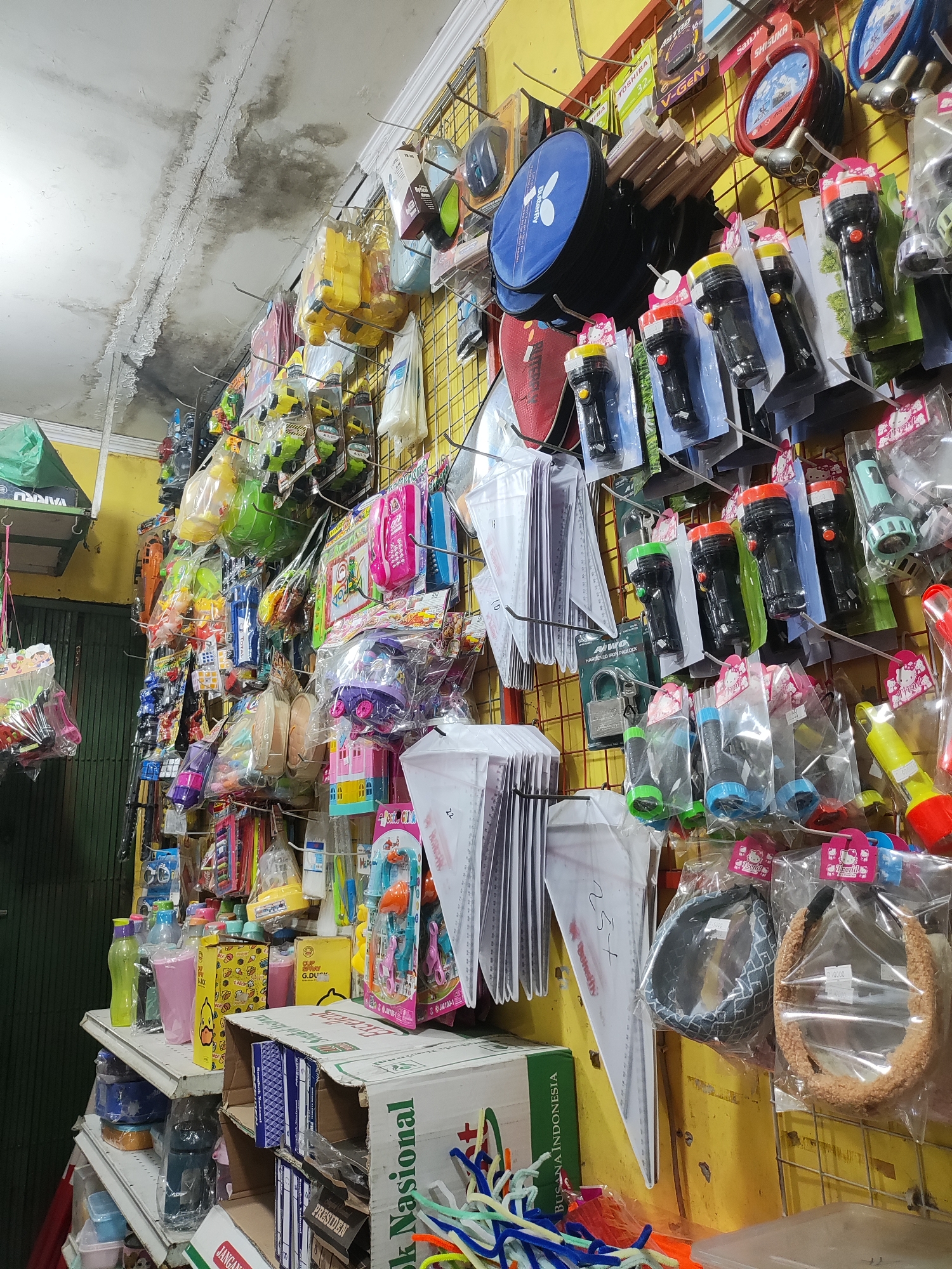 The image shows a wall-mounted display of assorted small items, likely for sale in a small shop or market stall in Wanajaya, Indonesia. The wall is painted yellow and a wire grid system is used for hanging the products. The items displayed include flashlights with varying colored caps, various toys packaged in plastic, a blue fabric bag, and some stationery items such as rulers and notebooks. There are also some headbands and a small mirror. The ceiling above shows signs of water damage. On shelves below the wall display, there are plastic bottles, boxes, and more packaged items. The overall impression is of a cluttered but organized retail environment.
