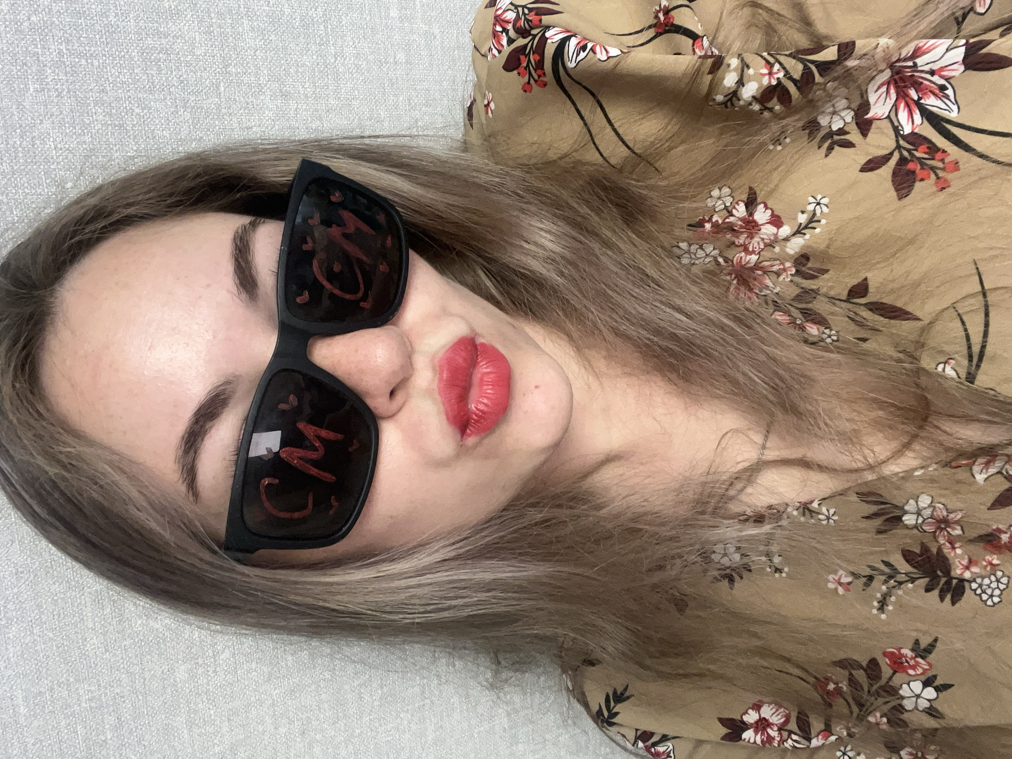 A woman with shoulder-length light brown hair is lying on a light grey textured surface. She is wearing sunglasses with the red letters "CM" written on each lens. She has light skin, distinct eyebrows, and red lipstick. She is wearing a floral-patterned blouse or dress in tan, white, and red. The image does not contain sufficient information to confirm the specific location in the city of Chaikovsky, Russia.