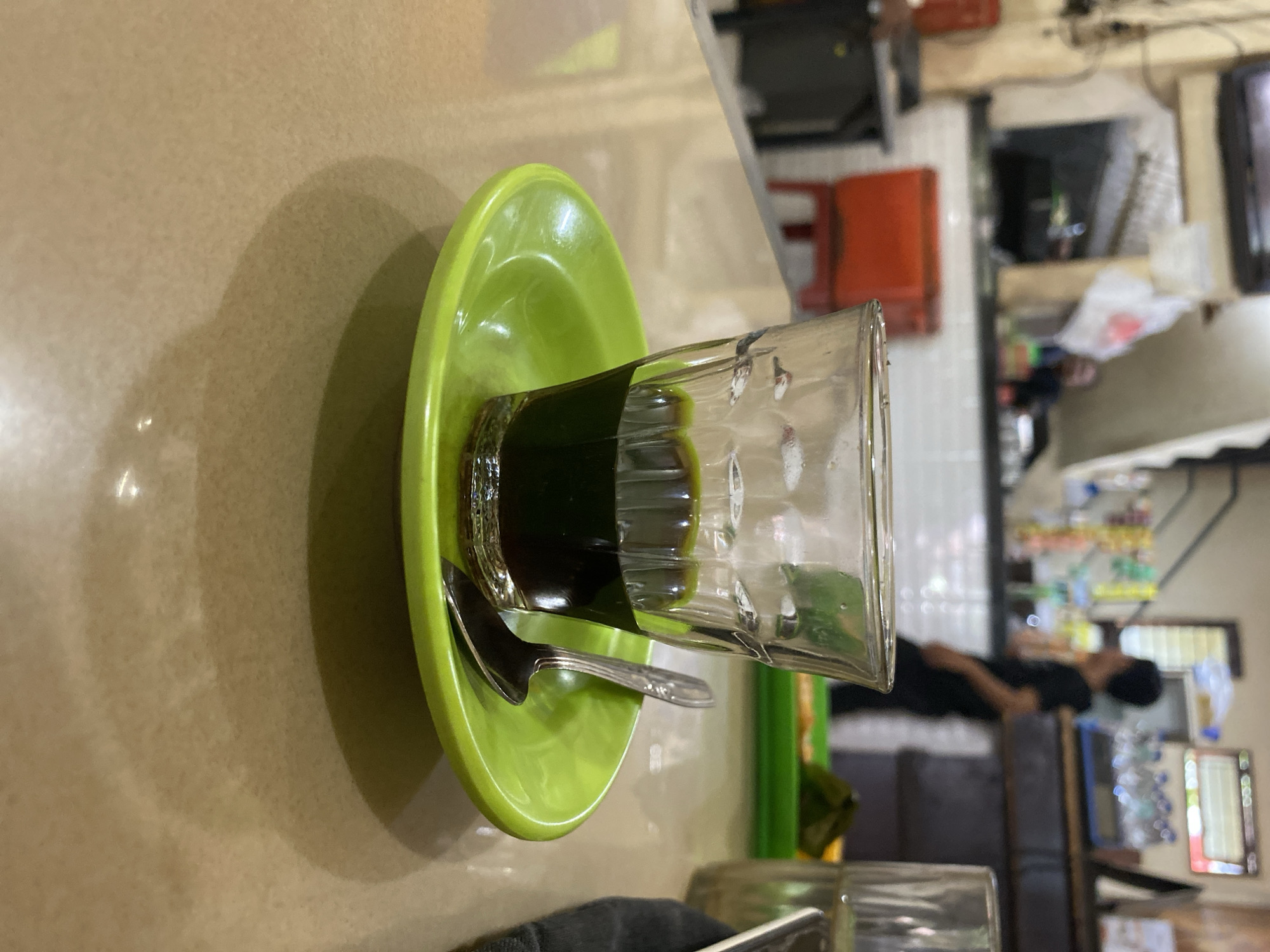 The image depicts a close-up view of a small glass filled with dark liquid, likely coffee, resting on a bright green saucer. A silver spoon is placed on the saucer, partially submerged in the liquid. The scene is set on a light brown, textured table. In the background, a blurred and out-of-focus environment suggests an indoor setting, possibly a cafe or eatery in Lhokseumawe, Indonesia. Elements such as a red chair, shelves with various items, and the silhouette of a person can be faintly discerned, indicating a public space. The lighting is subdued, creating shadows and highlighting the glass and saucer.