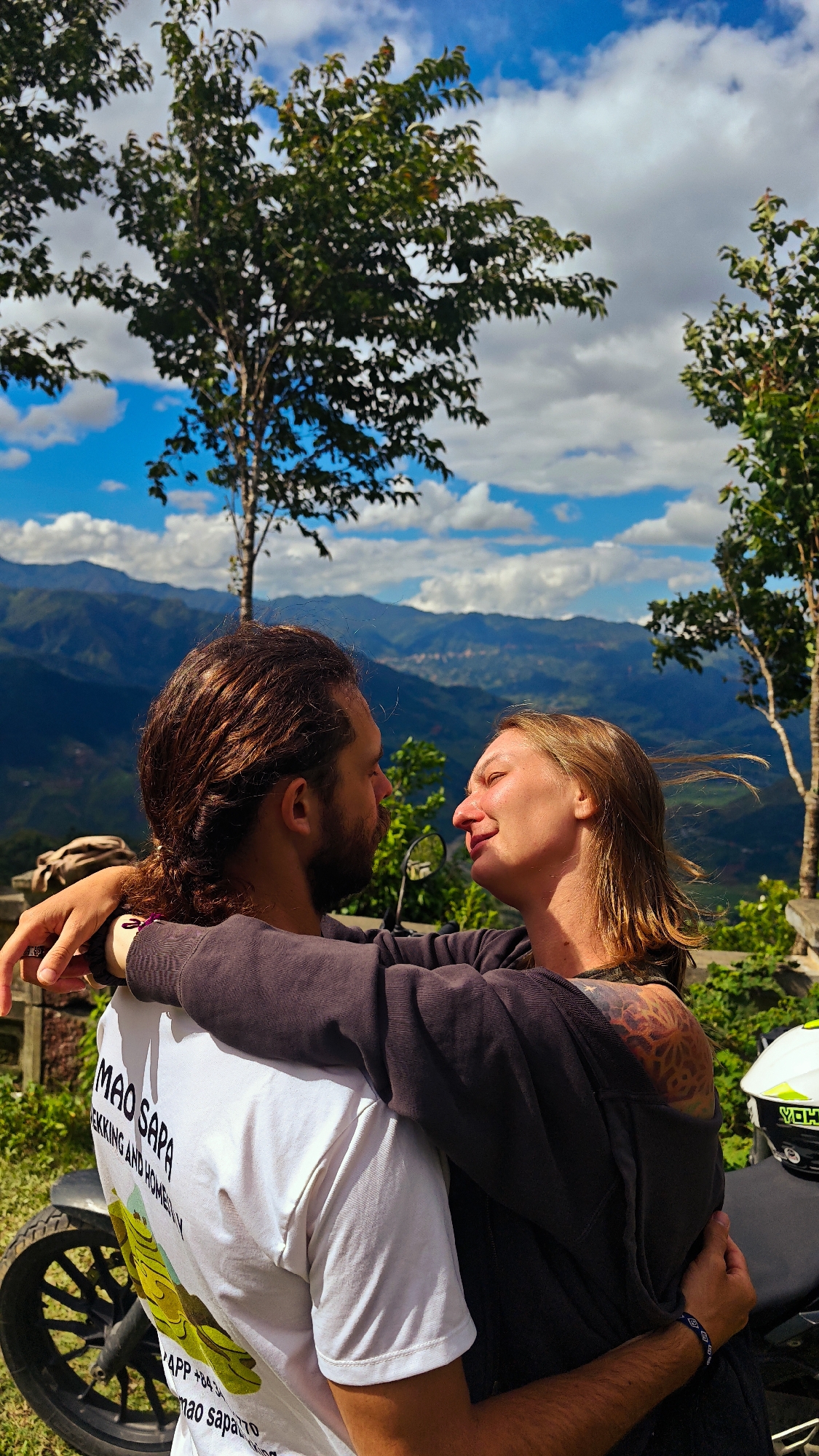 Two individuals, a male and a female, embrace at a scenic mountain viewpoint in Tu Le, Vietnam. The male, with long dark brown hair and a beard, is seen from the back-left, wearing a white t-shirt featuring "MAO SAPA TREKKING AND HOMESTAY" text and a yellow-green graphic. His arms are around the female's lower back. The female, positioned facing the male, is seen in profile with her eyes closed, a slight smile on her face, and light brown hair wind-swept to the right. She wears a dark grey long-sleeved top, revealing a geometric tattoo on her right shoulder and a blue patterned wristband on her left wrist. Her arms are wrapped around the male's shoulders/neck.
In the lower left, the front wheel and part of the frame of a black motorcycle are visible. A white motorcycle helmet with yellow-green accents is seen on the right, possibly resting on the motorcycle seat. A light-colored bag is visible on a stone railing in the mid-left background. The background features multiple layers of blue-hued mountains receding into the distance, with a distant settlement visible on one of the slopes. The sky above is bright blue with scattered white clouds. Green foliage and trees are present in the foreground and midground, framing the couple.