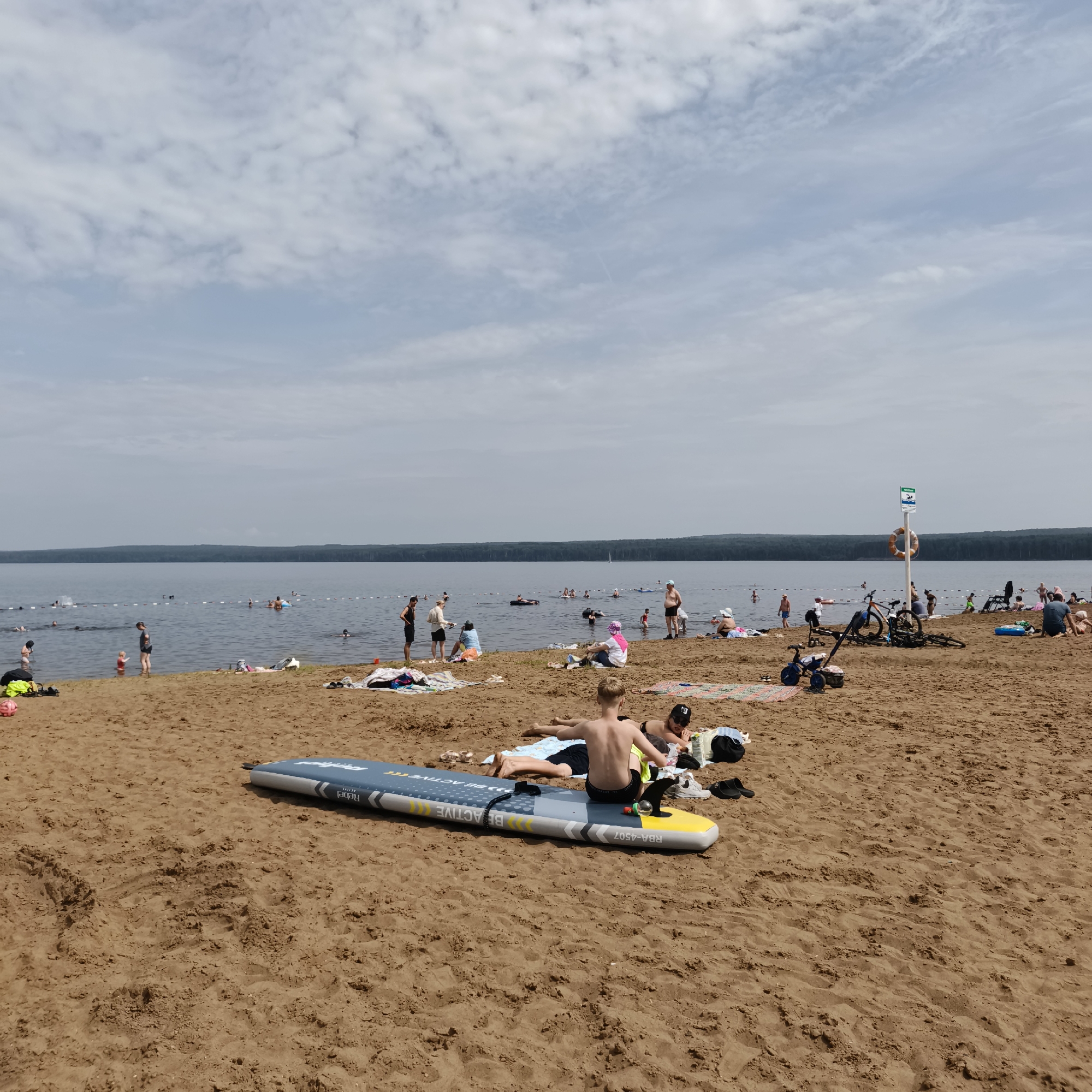 The image depicts a crowded sandy beach on a lake in Chaikovsky, Russia. In the foreground, two young males are lying on a blue and yellow inflatable paddleboard. One is sitting on the paddleboard, facing away from the camera, while the other is lying on his stomach. Nearby, several towels, shoes, and a pink ball are scattered on the sand.
Further back, numerous people are visible on the beach and in the water. Some are sunbathing on towels, while others are wading or swimming in the lake. A rope barrier divides the swimming area from the deeper water. Several bicycles and a small blue tricycle are parked on the beach, along with a white pole with a lifebuoy and a sign.
The lake stretches across the middle ground, with a dense line of trees forming the distant shoreline. The sky is partly cloudy, with white clouds scattered across a pale blue expanse. The overall scene is one of a busy summer day at a recreational lake.