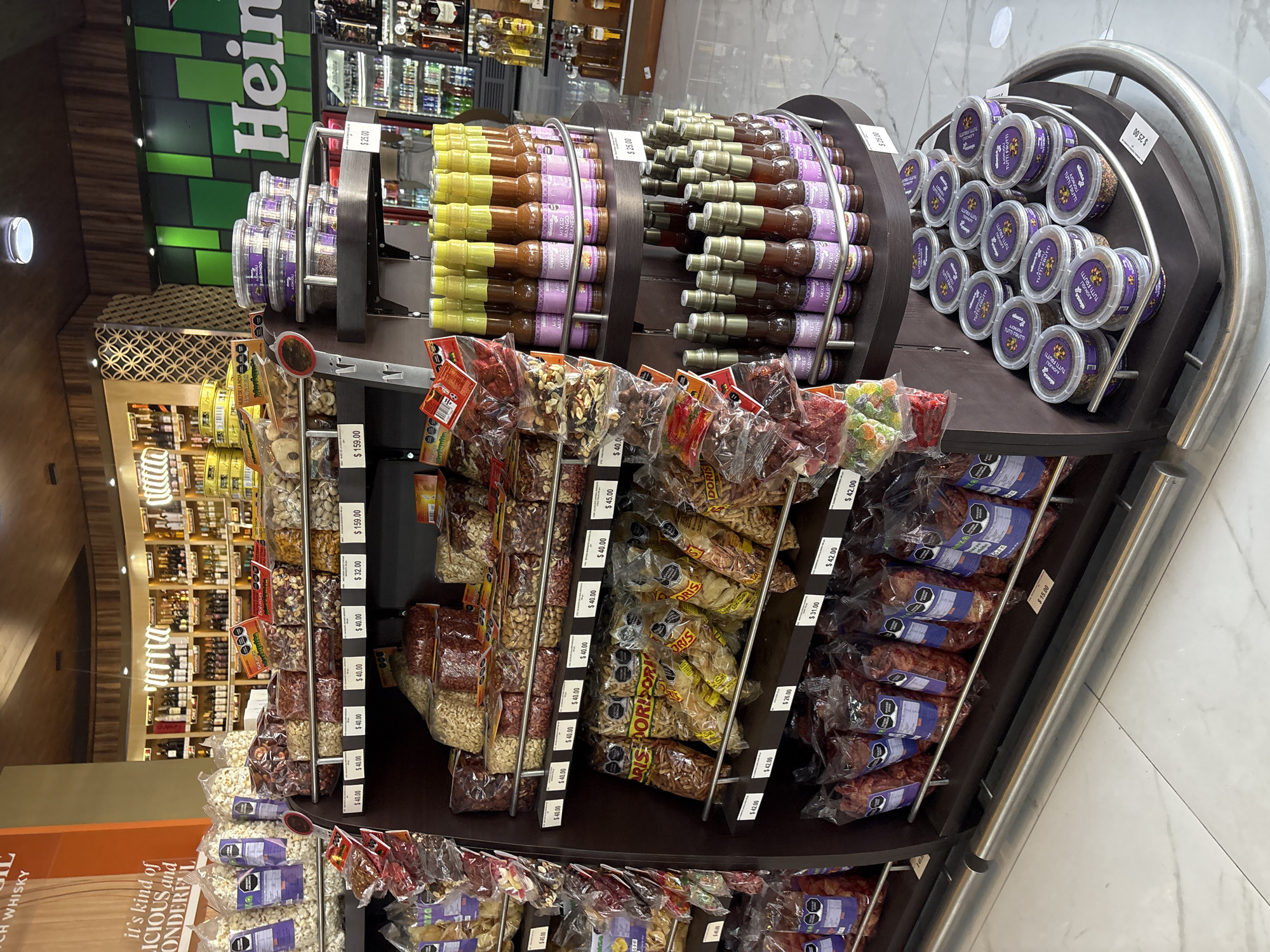 The image shows a store display rack filled with various food products. The top level features jars of what appears to be sauces or preserves in two rows and small plastic containers filled with food item in the row to the right. Below, the rack is stocked with a variety of packaged nuts, snacks, and other food items, each labeled with price tags. The prices are in Mexican pesos (MXN), some prices visible are $25.00, $15.00, $42.00, $26.00, $31.00, $40.00, $45.00, $32.00, $159.00. In the background, more store shelves are visible, stocked with alcoholic beverages. One can see that the shop sells beers (Heineken), whisky and other alcoholic beverages. Based on the price tag, the store is located in Mexico. The OCR data says the city of San Bernardino Tlaxcalancingo.