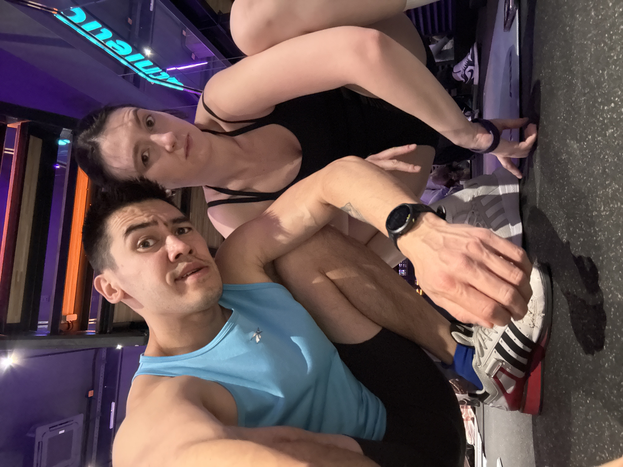 The image shows two people, a man and a woman, posing for a selfie in what appears to be a gym. The man is in the foreground, wearing a light blue tank top and black shorts. The woman is behind him, wearing a black sports bra and matching bottoms. Both are looking towards the camera. The background includes gym equipment and a reflective surface with lettering. The location is likely Kazan, Russia, based on the lettering on the reflective surface, which seems to be in Russian.