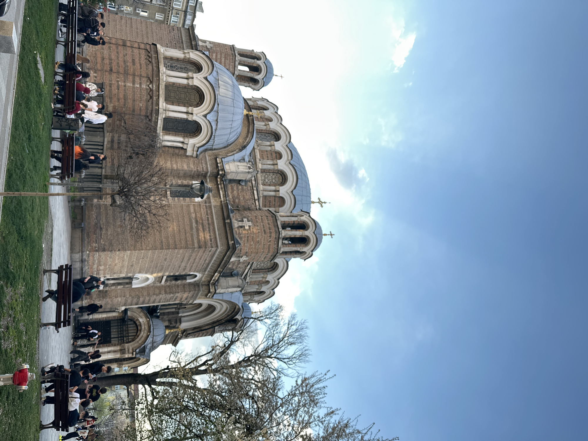 The image shows the St. Alexander Nevsky Cathedral in Sofia, Bulgaria. The cathedral is a large, ornate building with multiple domes topped with golden crosses. The sky is bright blue with some clouds. In the foreground, there are people sitting on benches along a walkway adjacent to the cathedral. The cathedral is made of light brown brick, and the domes are covered in a dark gray material. Bare trees stand in front of the cathedral, suggesting it's either late autumn or early spring.