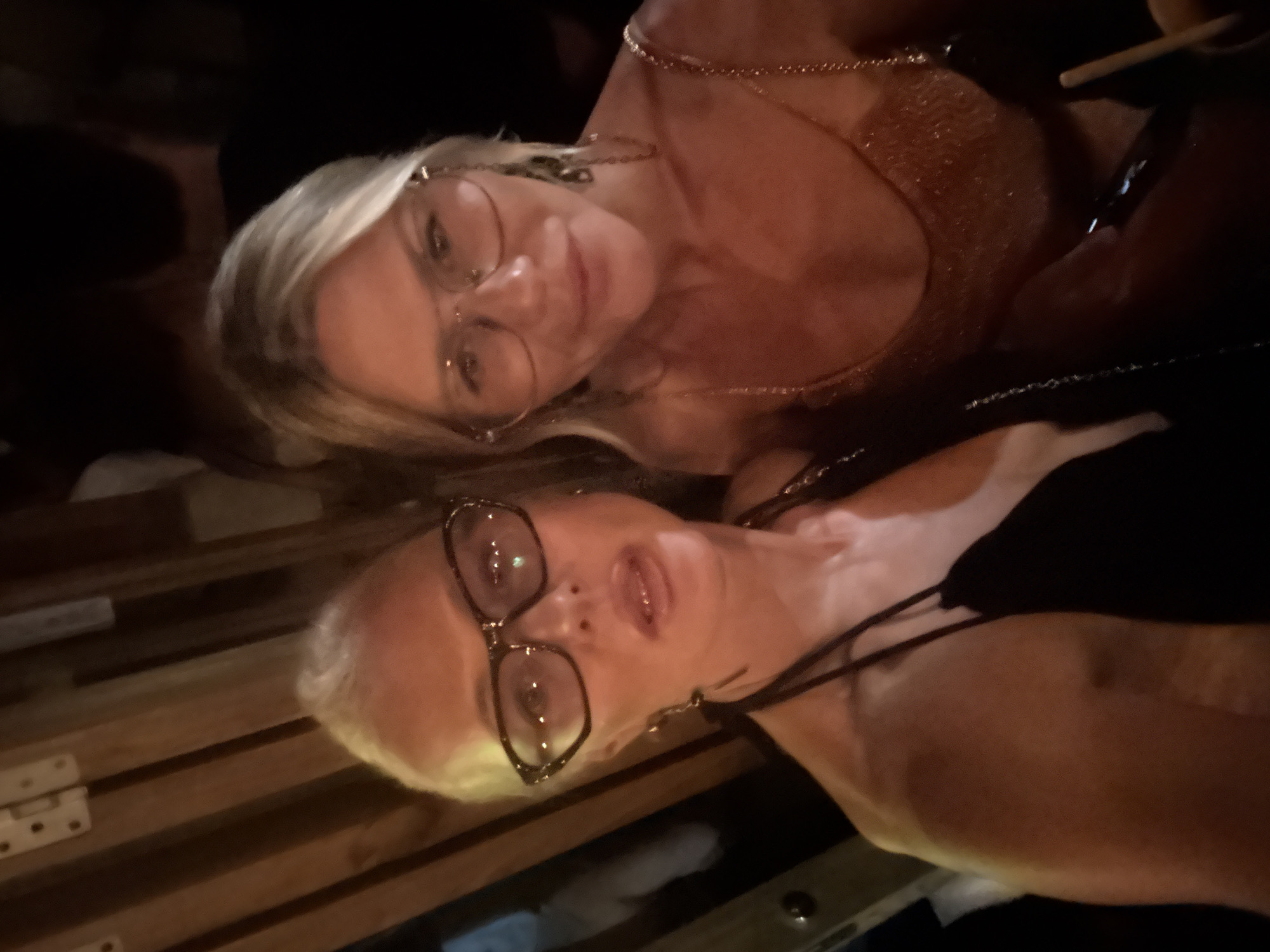 Two women are posing for a selfie in what appears to be a dimly lit indoor setting, possibly a bar or restaurant. The woman on the left has blonde hair, wears large, thick-rimmed glasses, and is looking slightly upward with her mouth open. She has on a black top with thin straps. The woman on the right also has blonde hair and wears aviator-style glasses. She is looking directly at the camera with a slight smile and is wearing a sparkly, possibly gold, top with thin chain straps. Both women are wearing jewelry. The background consists of wooden beams and is dark, suggesting low lighting conditions. The overall atmosphere is casual and social.