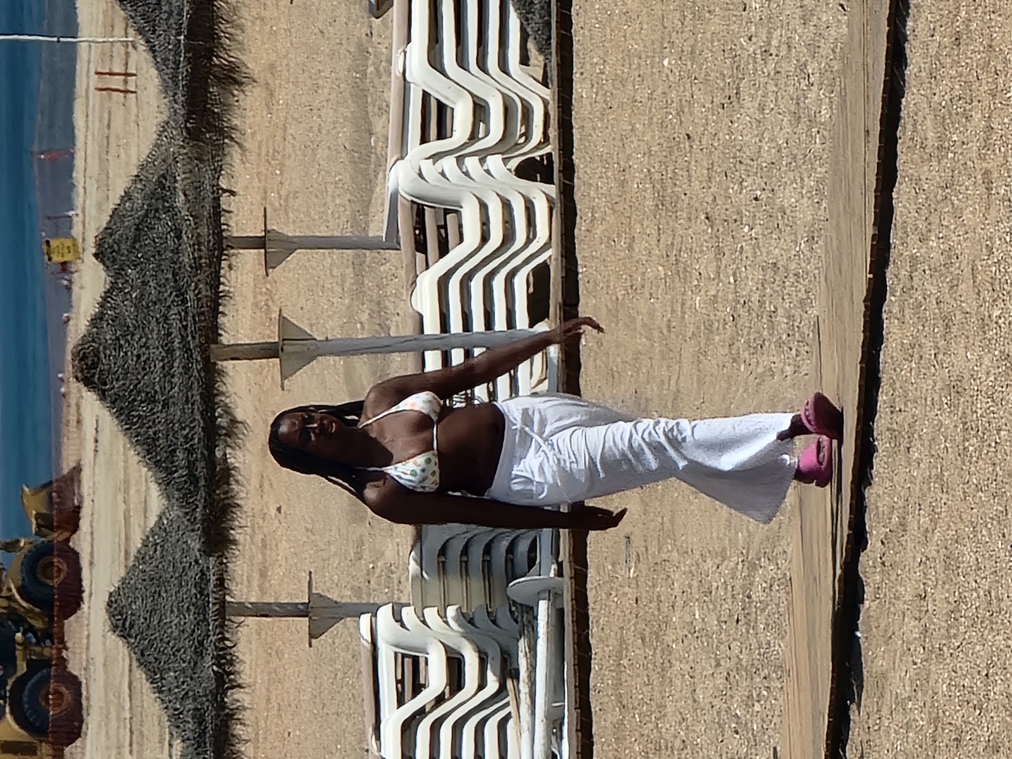 A young Black woman with long braids is walking on a wooden boardwalk on a sandy beach in Quarteira, Portugal. She is wearing a white bikini top with a polka dot pattern and loose white pants. She also has on bright pink slides. The beach is lined with rows of stacked white lounge chairs and thatched umbrellas. In the background, a yellow construction vehicle is visible on the sand near the water's edge. The sky is clear and blue, and the ocean is visible in the distance. The overall scene suggests a warm, sunny day at a popular tourist destination.